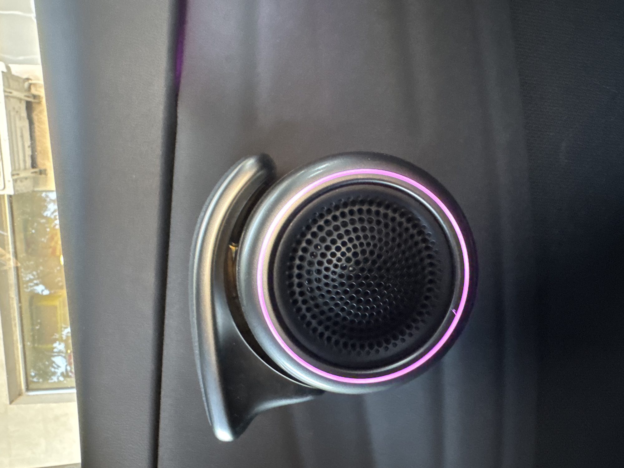 This is a close-up, eye-level shot of a car's interior door panel, specifically focusing on a speaker and a door handle. The speaker is circular with a perforated grille and is surrounded by a metallic ring that has a pinkish-purple ambient light emanating from behind it. Above the speaker, a metallic door handle curves over it. The surrounding door panel is made of a dark, textured material, possibly plastic or leather. The background shows a blurred reflection in what appears to be a window or a screen, with hints of outdoor scenery, possibly indicating the car is parked. There are no people or animals present in the image. The image provides no information about the city of Yerevan, Armenia.