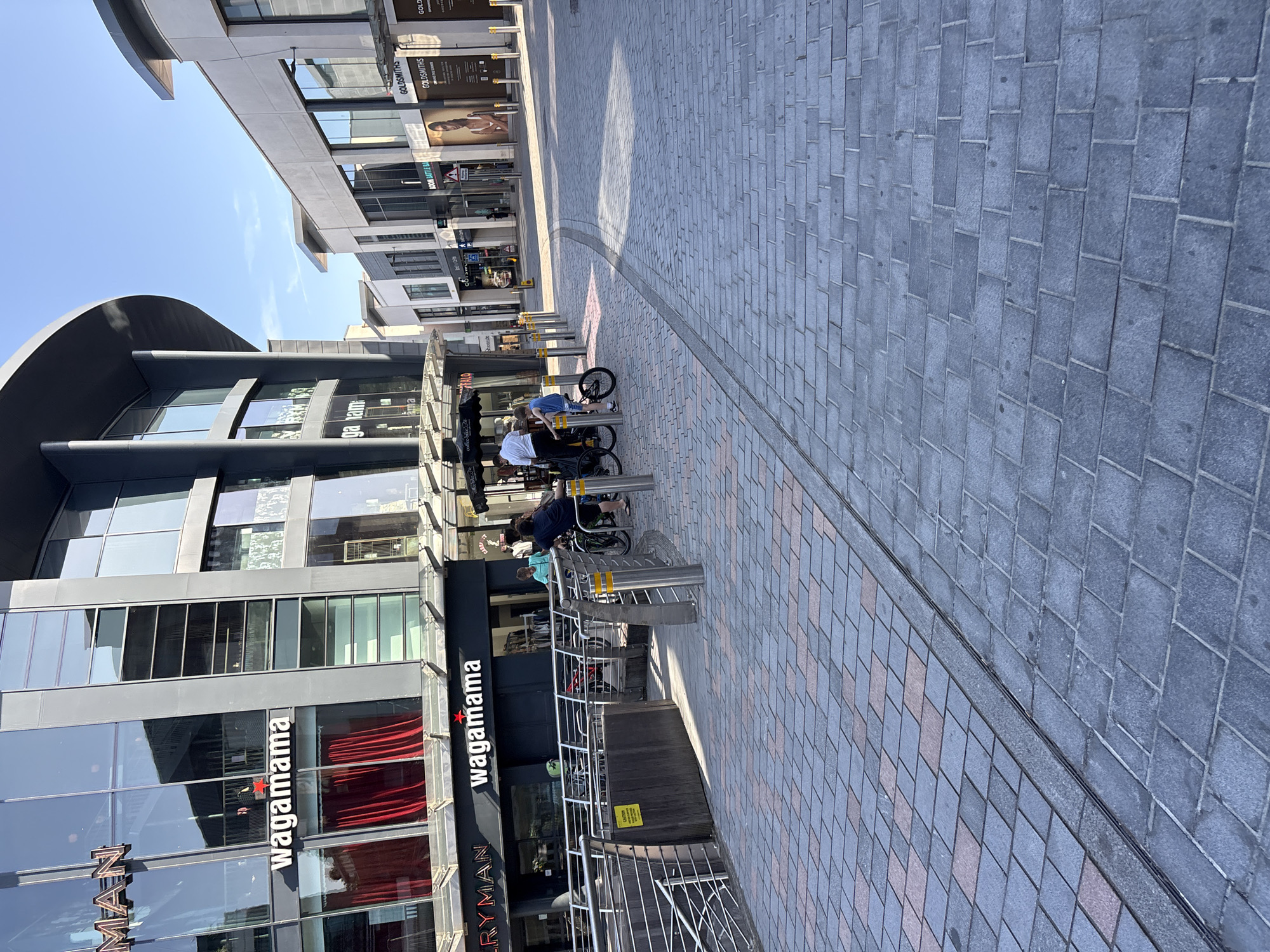 The image shows a sunny outdoor scene in the city of Chelmsford, United Kingdom, likely on a pedestrianized street or plaza. In the foreground, a patterned brick walkway leads the eye towards a building with a modern glass facade. Several shops are visible, including "Wagamama" with its distinctive red curtains.
A young boy is seen on a bicycle in the middle ground, with an adult nearby. Further back, other individuals are walking or standing, suggesting a public space with moderate foot traffic. The architecture is contemporary, with large windows and clean lines. The sky is clear and blue, indicating good weather. The overall impression is of a vibrant urban environment.