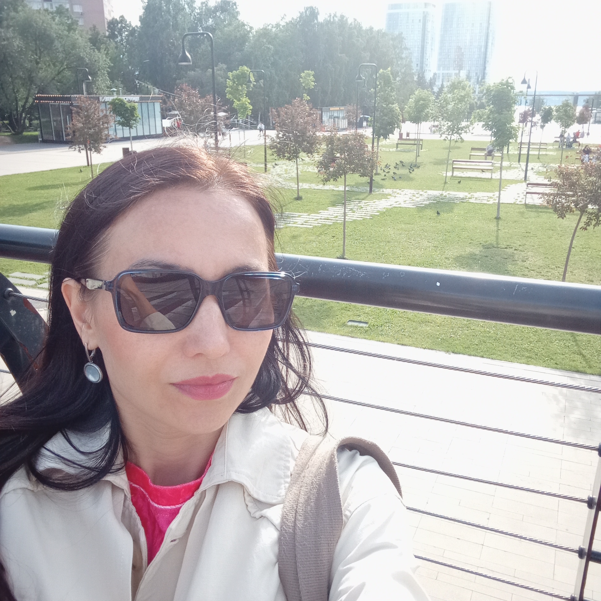This image depicts a woman taking a selfie in an urban park on a sunny day. The woman occupies the lower left portion of the frame, positioned in front of a black metal railing with multiple cable supports. She has dark hair, wears dark rectangular sunglasses, a light-colored open jacket over a pink top, and a light blue-grey oval earring on her left ear. Her lips are tinted with pink lipstick. The reflection in her right sunglass lens reveals a partial view of an arm or hand and the reversed word "balencia" (likely a brand name).
Behind the railing, the scene unfolds into a well-maintained park featuring green grass, numerous small trees, and pathways constructed from light-colored rectangular paving slabs. Several pigeons are scattered across the grass areas. Modern street lamps with dark fixtures are intermittently placed throughout the park. In the middle ground, a contemporary glass-fronted building with a dark, possibly corrugated, roof is visible among the trees on the left. Further in the background, to the far left, a multi-story residential building is partially visible. Two prominent, tall, modern high-rise buildings with numerous windows dominate the distant skyline on the right side of the frame. Park benches are scattered throughout the grassy areas.
This specific location is identified as the Central Embankment (Центральная набережная) or an adjacent area within Kirov Park (Парк им. Кирова) in Izhevsk, Russia, overlooking the Izhevsk Pond. The distinctive modern park design, the glass kiosk, and the specific high-rise buildings in the background are characteristic features of this renovated urban area in Izhevsk.