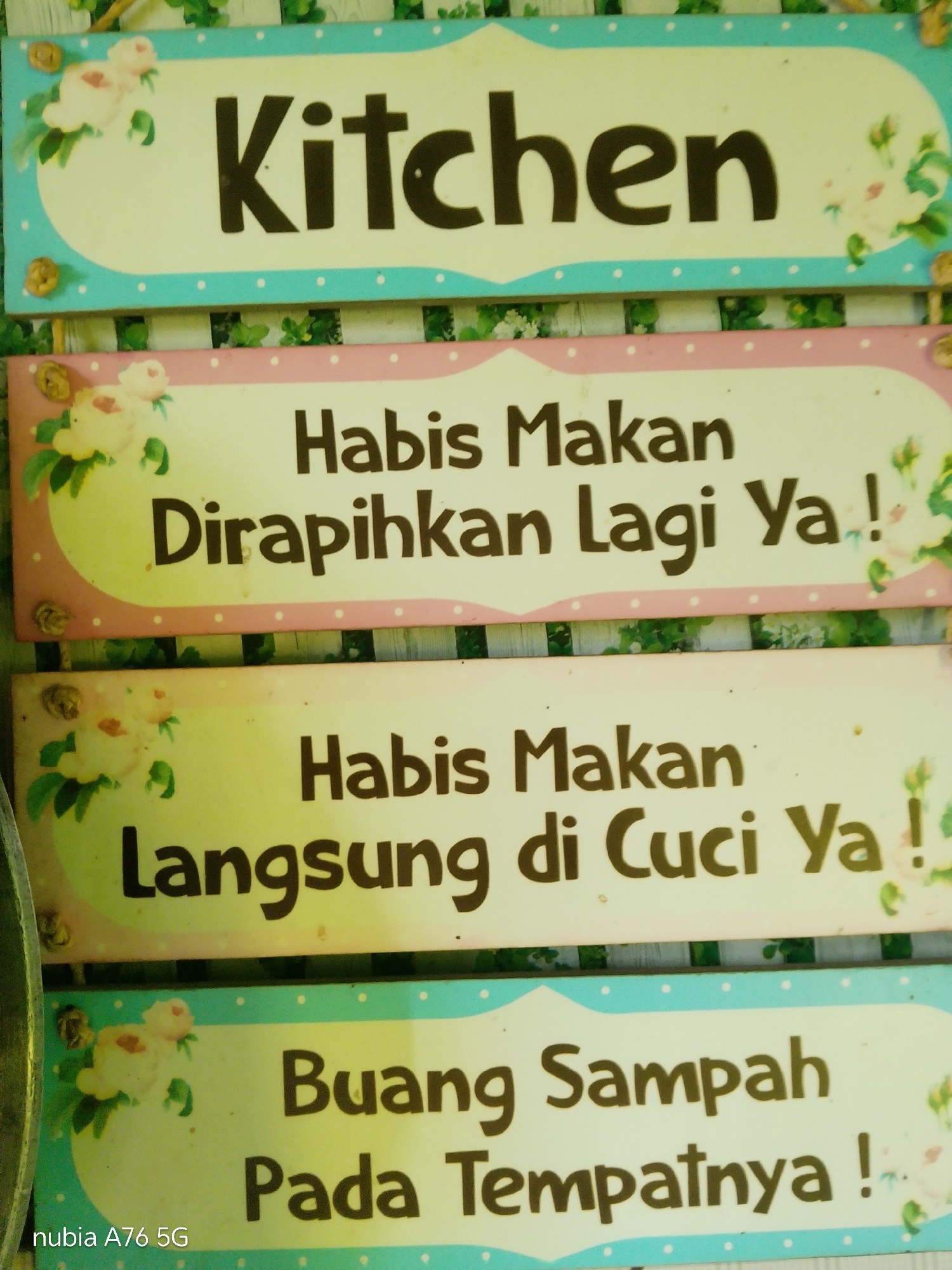 The image displays four signs with Indonesian text and floral decorations, likely intended as instructions or labels. The top sign reads "Kitchen". The second sign says "Habis Makan Dirapihkan Lagi Ya!" which translates to "After Eating Tidy Up Again!". The third sign states "Habis Makan Langsung di Cuci Ya!" meaning "After Eating Wash It Right Away!". The bottom sign reads "Buang Sampah Pada Tempatnya!" or "Throw Trash In Its Place!". The signs have a pale yellow background with decorative borders and floral motifs in pink and light blue. The image also contains the text "nubia A76 5G" in the bottom left corner, which is likely a watermark from the device used to capture the photo. The background appears to be a wooden surface, possibly a wall or fence, with some greenery visible between the signs. There is no visible indication of the location being in Cakung, Indonesia.