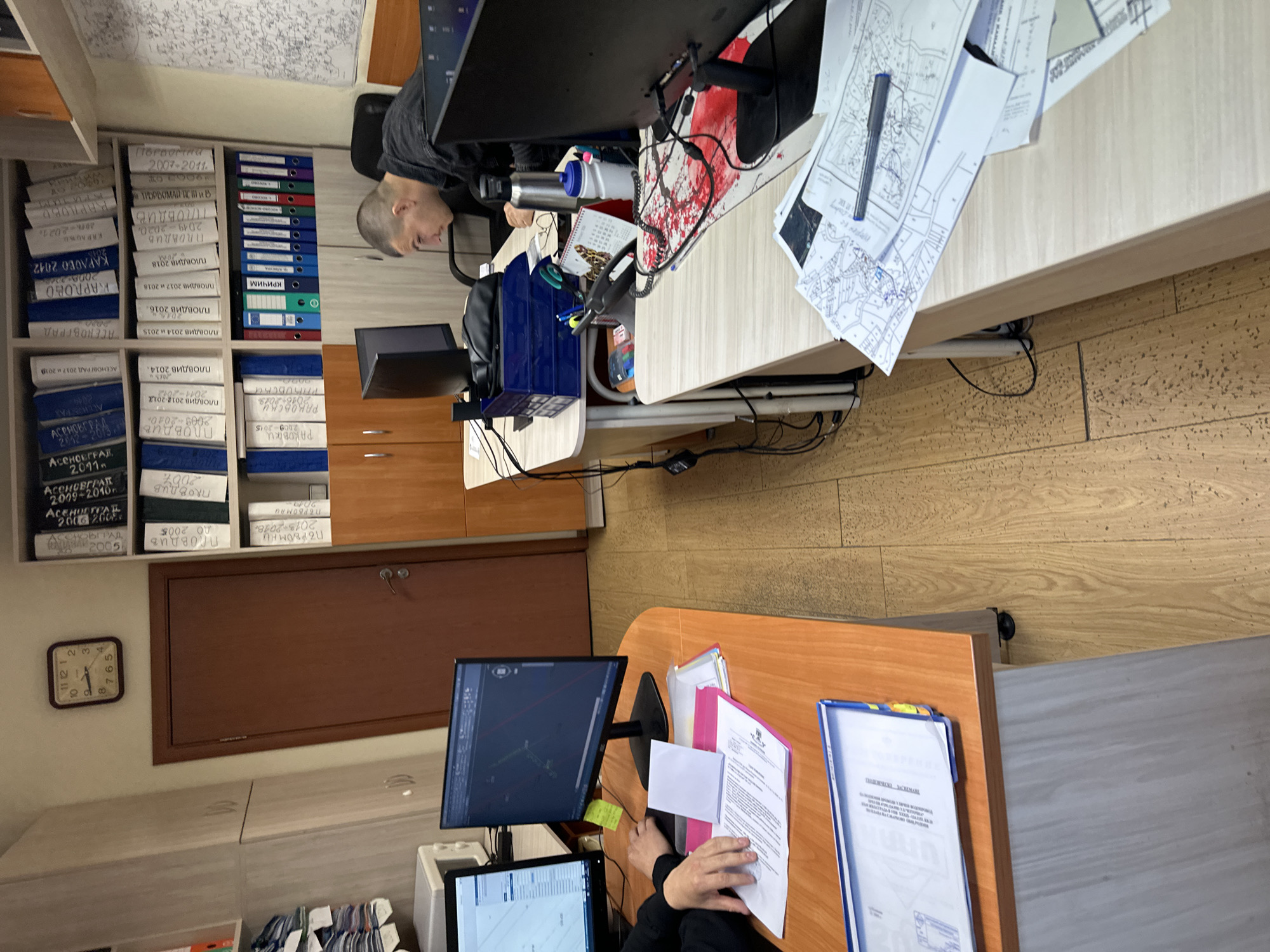 The image shows an office setting with two desks. On the left, there's a tall bookshelf filled with labeled folders, many referencing "Plovdiv", "Asenovgrad", "Karlovo", and "Pervomay" along with years from the early 2000s to the late 2010s. A clock on the wall displays 10:55. A person with a shaved head sits at a desk facing away from the camera, working on a computer. There's also a desk on the right where someone is reviewing papers. The general environment seems to be a documentation or administrative office. The labels on the folders and the references to specific towns (Plovdiv, Asenovgrad, Karlovo, and Pervomay) suggest a local or regional scope of work. Given that Plovdiv is a major city in Bulgaria, the office might be involved with administrative or legal matters pertaining to properties, permits, or other local government affairs.