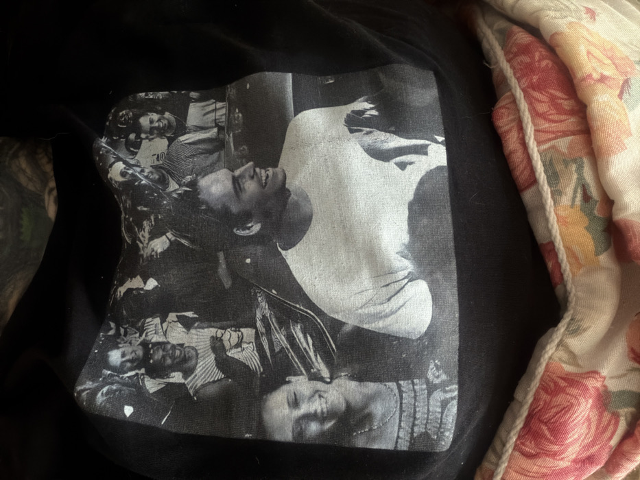 A black t-shirt lays on a surface, featuring a black and white print of a man in a white shirt surrounded by a crowd. The print has a vintage, slightly faded look. To the right of the shirt, a floral-patterned fabric with roses and a neutral-colored border lies crumpled. The photo is somewhat close-up, obscuring the surrounding environment.