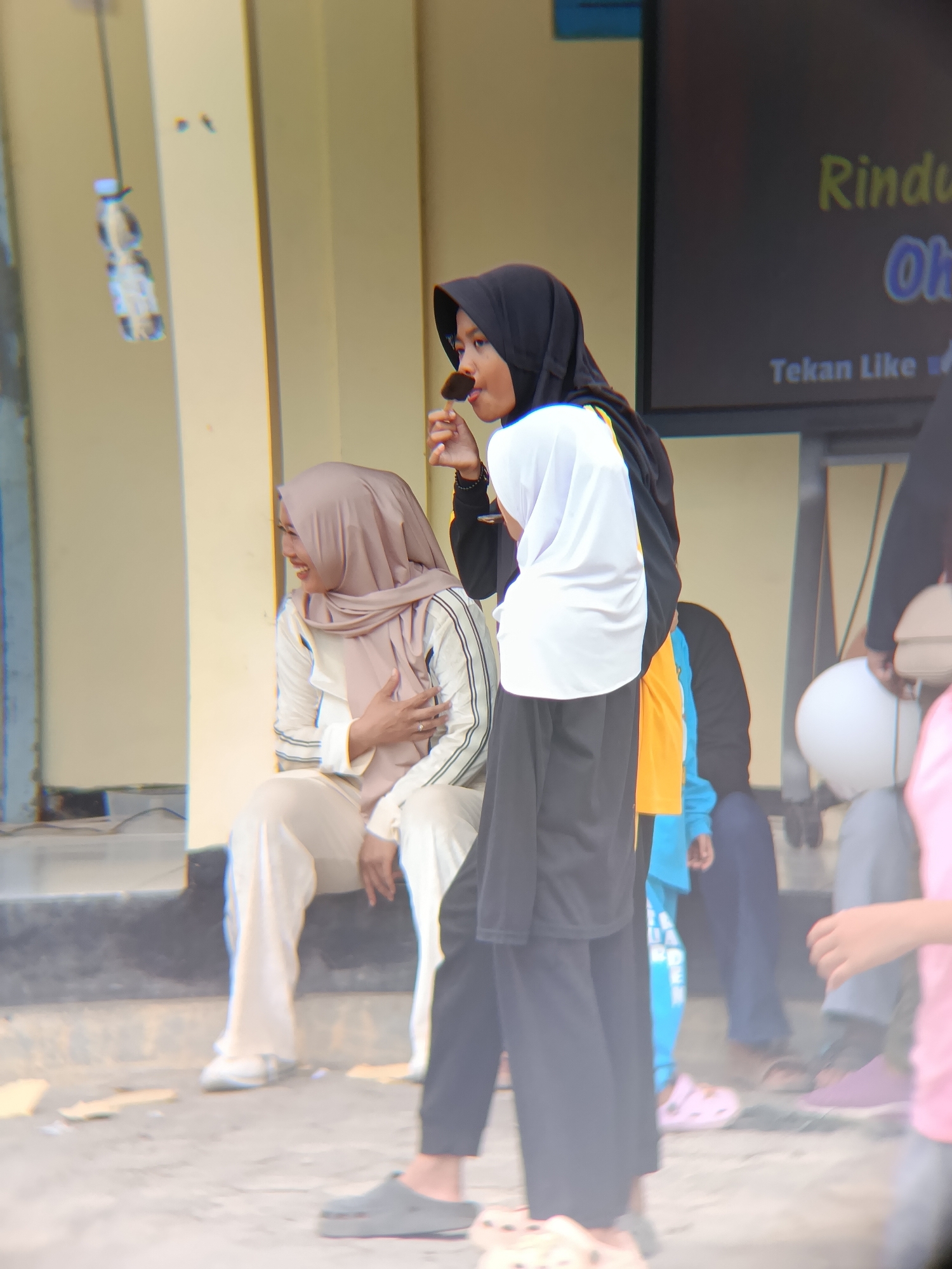 In the city of Bulaksari, Indonesia, a group of people are gathered outdoors. The scene appears to be a casual event, possibly a community gathering or celebration. In the foreground, a woman wearing a black hijab and a white and black outfit is holding a piece of food on a stick to her mouth, appearing to be eating. To her left, another woman is seated, wearing a beige hijab and a matching outfit. She is looking to her left and laughing, with her hand on her chest.

Behind them, other individuals are visible, some of whom are partially obscured. One person is wearing a bright orange shirt and another a blue tracksuit with white lettering. Several pairs of feet are visible, shod in various footwear including pink Crocs, brown sandals, and grey slip-on shoes.

The environment consists of a light-colored building with vertical columns. In the background, a dark screen displays text in yellow and blue, with the words "Tekan Like" visible, suggesting it is a digital display or monitor. A clear plastic bottle hangs from a string in the upper left portion of the image. The ground is rough and appears to be paved with concrete or stone. The overall atmosphere seems lively and relaxed.