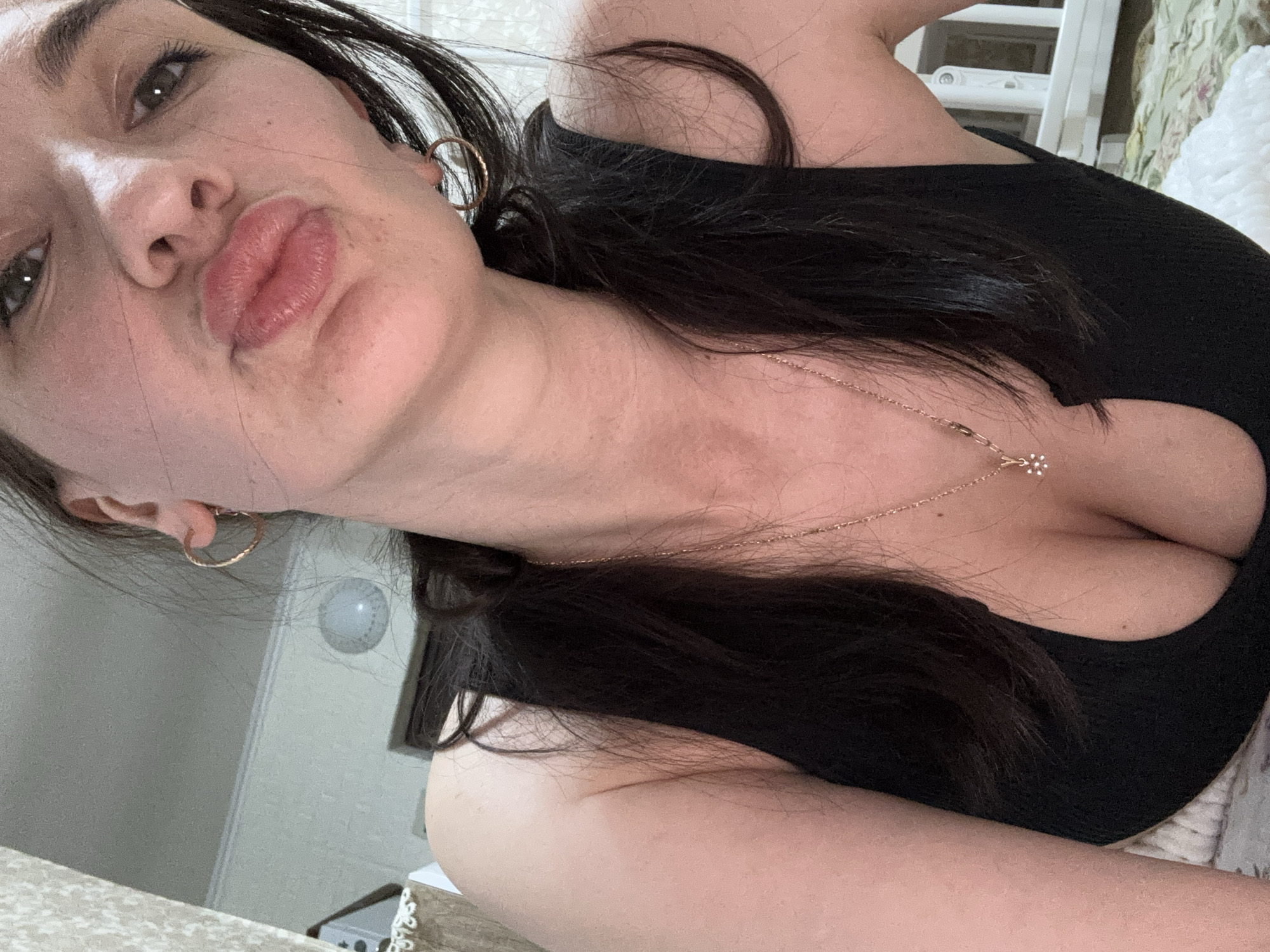 A close-up, selfie-style image of a woman with fair skin, dark hair, and light eyes is presented. She is wearing gold hoop earrings and a delicate gold necklace with a small flower-shaped pendant. The woman is wearing a black tank top with a low neckline. Her lips are pursed. The background is indistinct, but suggests an interior setting with a white wall and a glimpse of a white ladder-like structure. There is a light fixture visible on the wall. The image does not contain information to determine the location as Sochi, Russia.