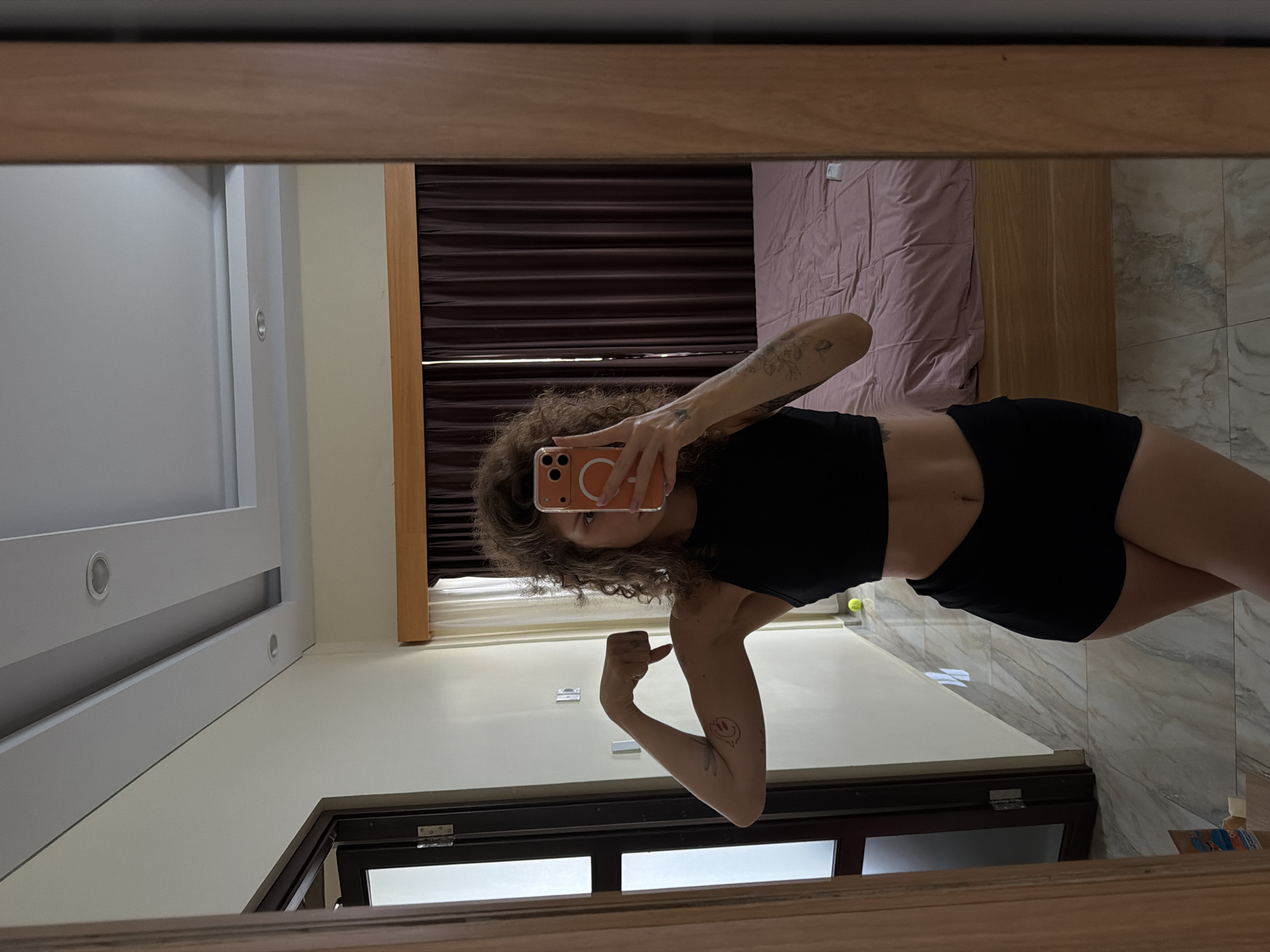 The original image, which is rotated 90 degrees clockwise, shows a person with curly brown hair taking a mirror selfie. The individual, wearing a black crop top and black shorts, holds an orange smartphone, which partially obscures their face. They are flexing their left arm, revealing tattoos on both arms; one on the left forearm depicts floral or foliage patterns, and another on the right bicep features a distinctive, possibly smiling, design.
The reflected background reveals an indoor setting, likely a bedroom. The room features light cream-colored walls and a light-toned tiled floor, resembling marble or stone. Behind the person, a bed with a light pink duvet is visible, along with dark brown horizontal window blinds. Recessed spotlights are embedded in the ceiling. The mirror itself is framed by light brown wood. A tennis ball is visible on the floor near the person's right leg. The bright indoor lighting suggests it is daytime. The selfie captures a confident, casual pose. The setting is contextually located in North Kuta, Indonesia. No explicit text is visibly legible within the image.