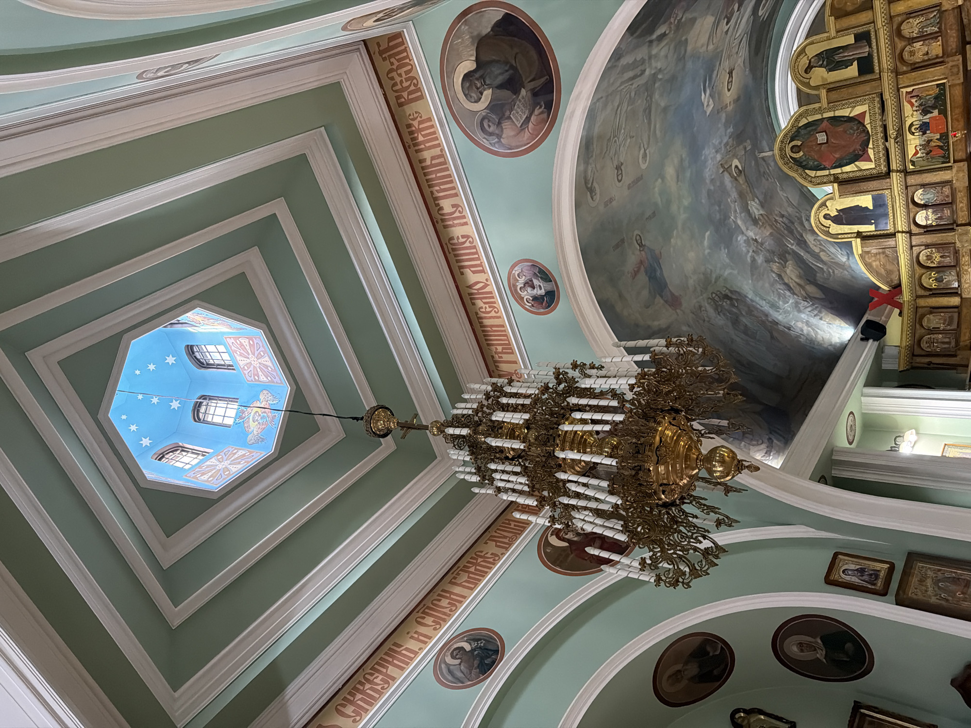 This is an interior shot of a church, likely Orthodox given the iconography. The scene is captured from a low angle, looking upwards towards the ceiling and a central octagonal skylight. The walls and ceiling are painted a soft green with ornate white moldings creating a tiered effect, drawing the eye towards the skylight.
The octagonal skylight features a bright blue ceiling adorned with white stars, and windows on its upper facets. Below the skylight, a large, ornate brass chandelier hangs, its numerous candle-like fixtures gleaming.
The ceiling is further decorated with religious paintings and iconography. One large mural depicts a cloudy, heavenly scene with figures. Along the walls, circular icons of saints or religious figures are visible, and a section of text in Cyrillic script is painted onto the wall. The overall impression is one of grandeur and spiritual atmosphere. The lighting appears to be a mix of natural light from the skylight and interior lighting, creating a somewhat dramatic effect.
The location is identified as Sochi, Russia. There is no indication of the time of day, weather, or any specific activity or event taking place, but the setting suggests a place of worship. The emotions evoked are likely reverence and awe due to the architectural and artistic elements.