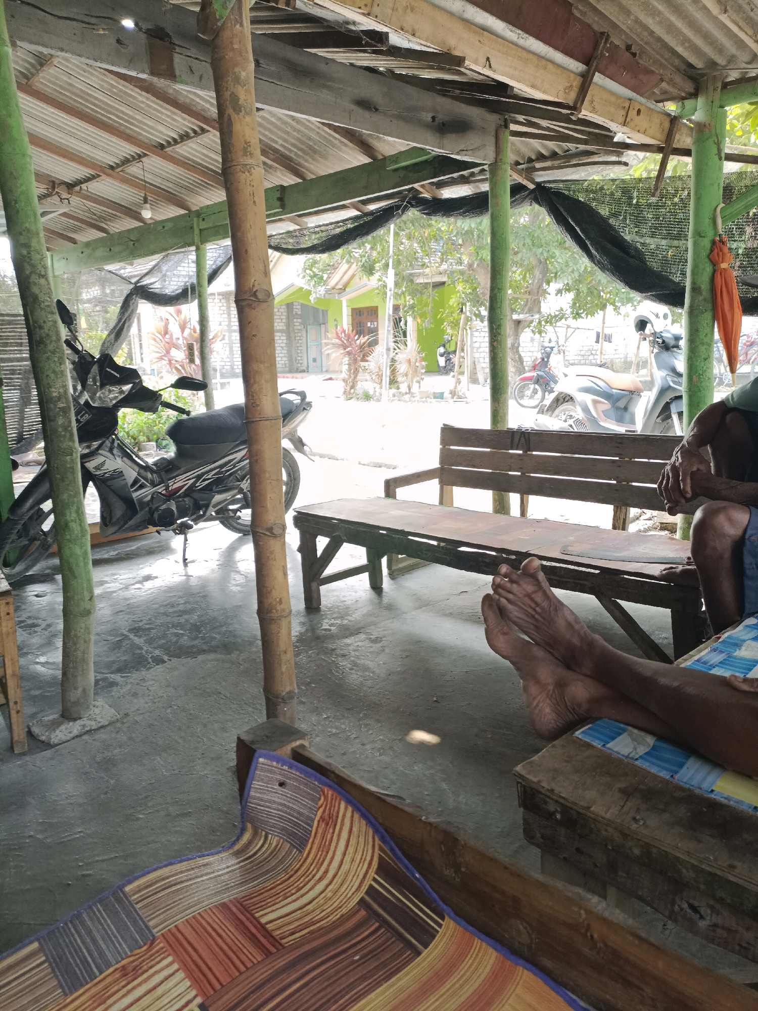 The image depicts an outdoor seating area with a corrugated metal roof supported by green painted posts and bamboo poles. A motorbike is parked on the left side of the frame. In the mid-ground, a wooden bench runs horizontally. In the foreground, a person's bare feet are visible, resting on a wooden surface covered with a mat. The background reveals houses and trees, suggesting a residential or semi-rural setting, potentially in Rancang Kencono, Indonesia. The overall scene appears casual and relaxed.