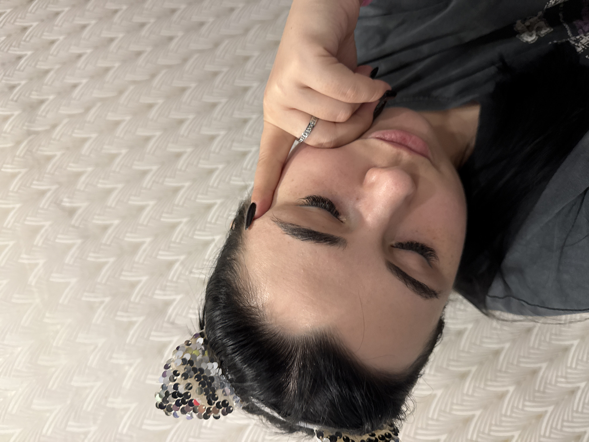 A woman with dark hair and black nail polish lies on a white, patterned surface. She is wearing a silver ring and a sparkly headband with cat ears. Her eyes are closed. She is touching her face with her hand. The location in the city of Chaikovsky, Russia is unknown.