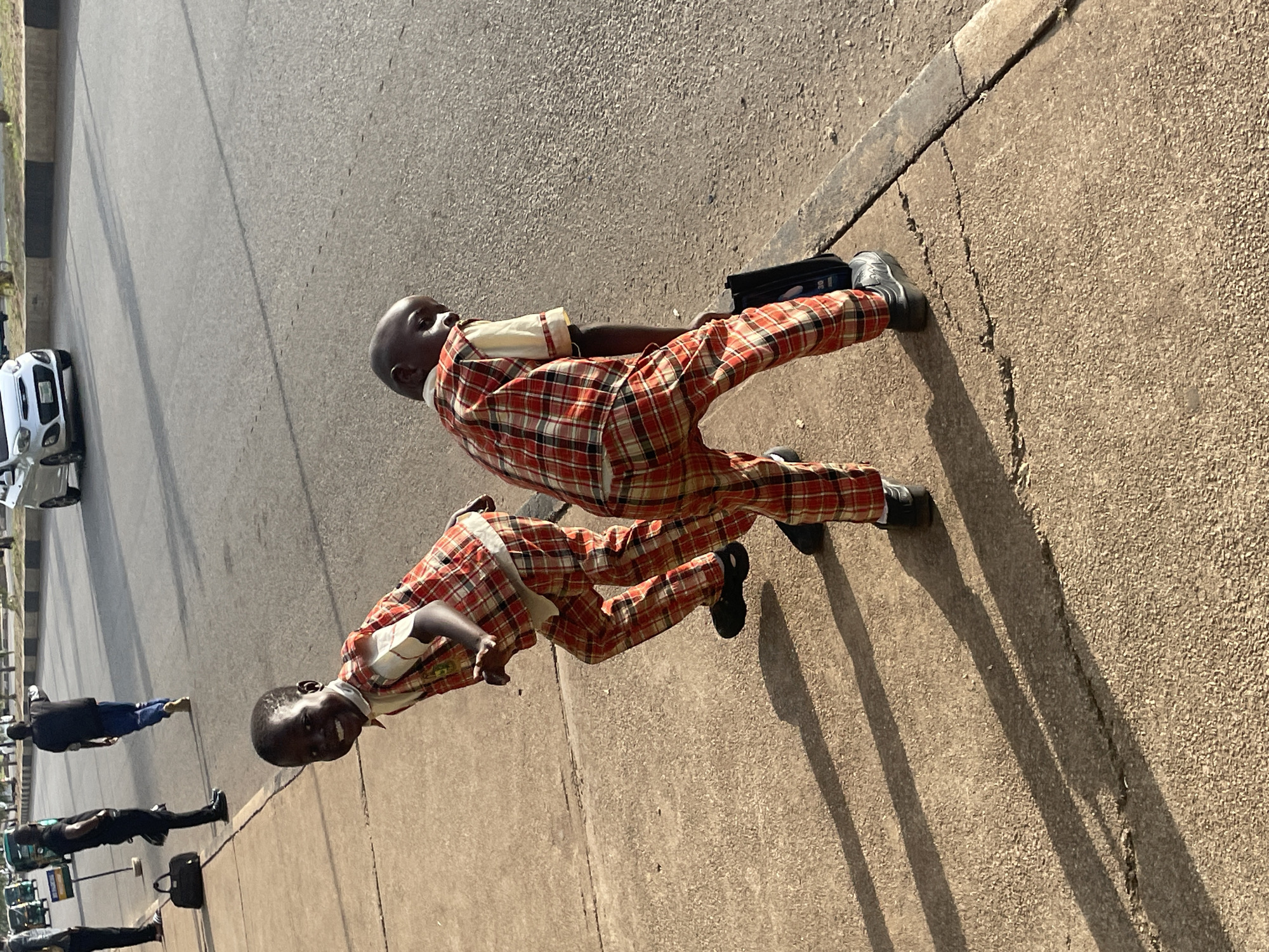 Two young boys wearing matching red and white plaid outfits, consisting of shirts and pants, are standing on a concrete sidewalk next to a paved road. The boy on the left is closer to the camera and looking directly at it, with a wide smile. He is wearing a white shirt with a red plaid vest and matching plaid pants. The boy on the right is facing away from the camera, bent over, holding a black bag or briefcase in his right hand. He is also wearing a similar plaid outfit. Their shadows are cast long on the sidewalk, indicating it is likely late afternoon. In the background, traffic can be seen on the road, including cars and possibly a bus. The environment appears to be an urban street in Jos, Nigeria, given the context provided. The road has distinct lane markings and a curb separating it from the sidewalk.