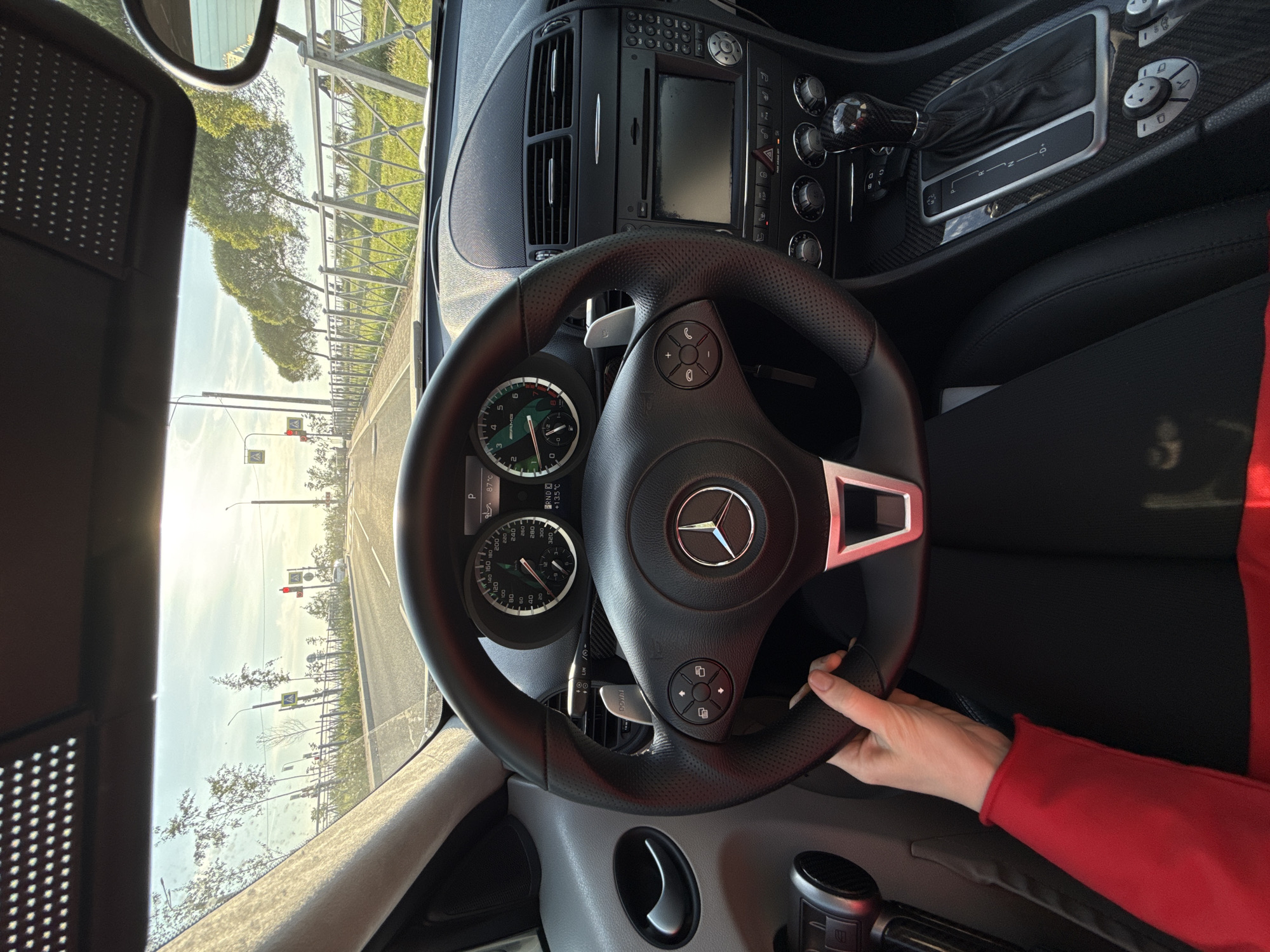 The image, rotated vertically, provides a first-person view from the driver's seat of a Mercedes-Benz car, likely in Saint Petersburg, Russia. The primary subjects are the car's interior, a person's hand, and the exterior road scene.
A person's hand, covered by a bright red sleeve, is holding the black, perforated leather steering wheel. The steering wheel features a prominent chrome Mercedes-Benz logo in the center and control buttons for media and phone functions. Directly behind the wheel, the instrument cluster is visible, displaying a speedometer marked up to "320" with "AMG" text, a tachometer, and digital readings for engine temperature ("87°C") and ambient temperature ("+13.5°C").
The dashboard is equipped with air vents, an infotainment system, and numerous control knobs and buttons. The gear selector, positioned to the right of the steering wheel and surrounded by a carbon fiber-like trim, shows the gear positions "P R N D".
Through the windshield, a bright, clear daytime scene is visible. A road extends into the distance, lined with green trees and a metal fence. A red traffic light is visible further down the road, suggesting the car might be stopped or slowing down. The sky is bright with some scattered clouds, indicating sunny weather, possibly during the morning or late afternoon due to the sun's angle.