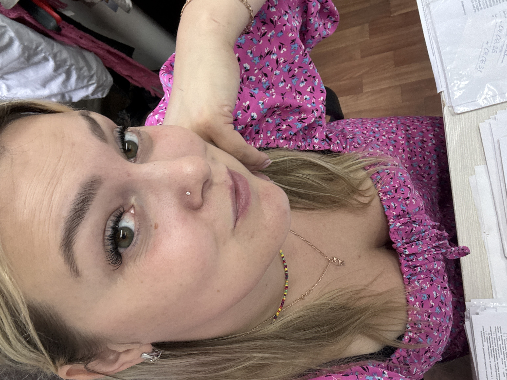 A woman with blonde hair, fair skin, and green eyes is shown in a close-up, high-angle shot. She is wearing a pink floral print dress with a low neckline and a black undergarment. She has long, false eyelashes and a nose piercing. She is wearing a beaded necklace and a pendant necklace. Her right arm is resting on her cheek, and her left hand is partially visible. The background shows a blurred office environment with wooden flooring and stacks of papers. The image does not contain any information about the location in the city of Noviy, Russia.