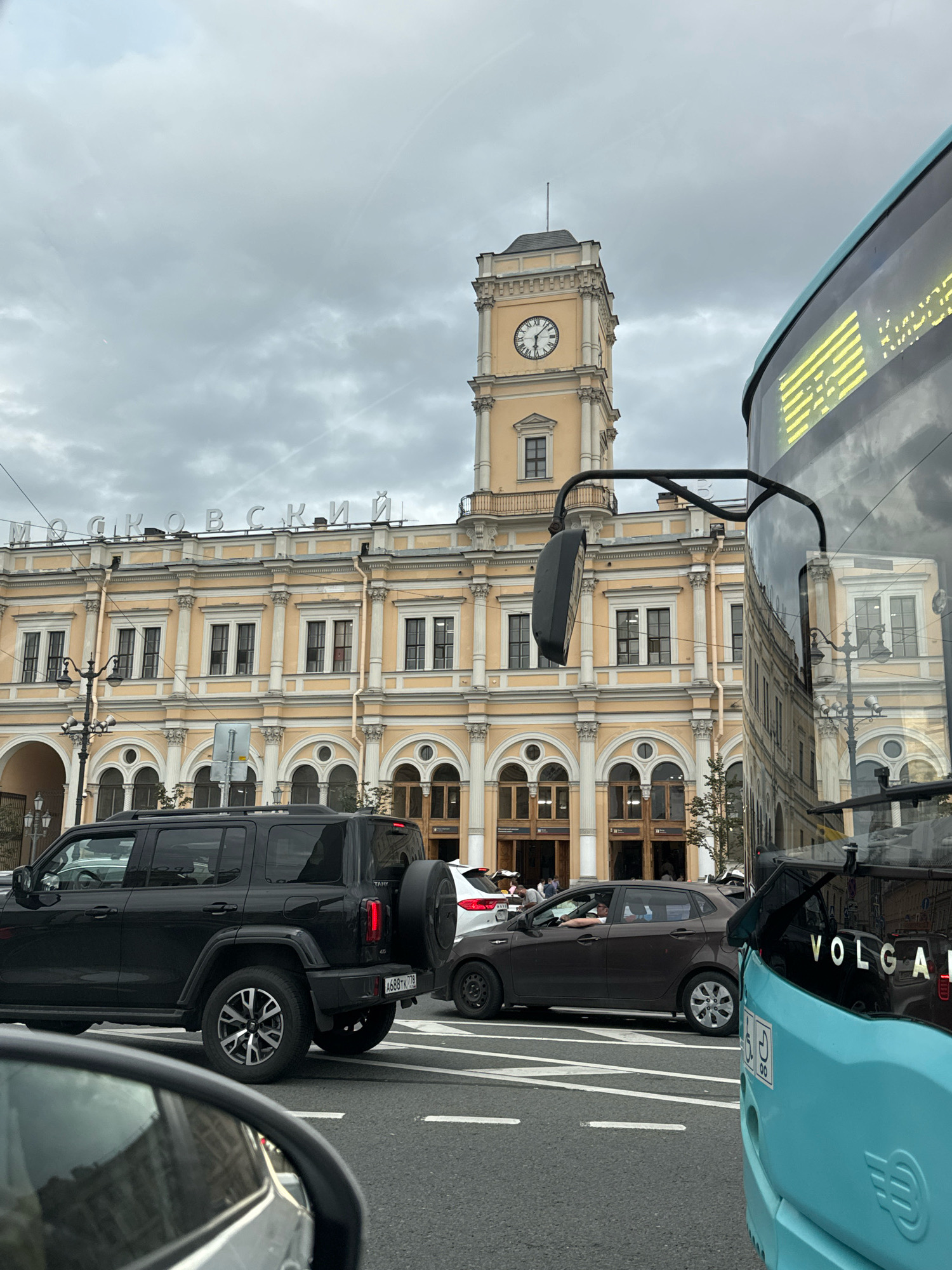 The image depicts a street scene in Saint Petersburg, Russia, with the iconic Moskovsky Railway Station as the prominent backdrop. The station, with its distinctive yellow facade and classical architecture, features a tall clock tower displaying Roman numerals. The sky is overcast, suggesting a cloudy day. In the foreground, traffic is present, with several vehicles visible. A black SUV is parked on the right side of the frame, while a light blue bus, with "VOLGA" written on its side, occupies the right portion of the image. A brown car is seen driving on the road. The scene captures the bustling atmosphere of a city intersection with a significant historical landmark.