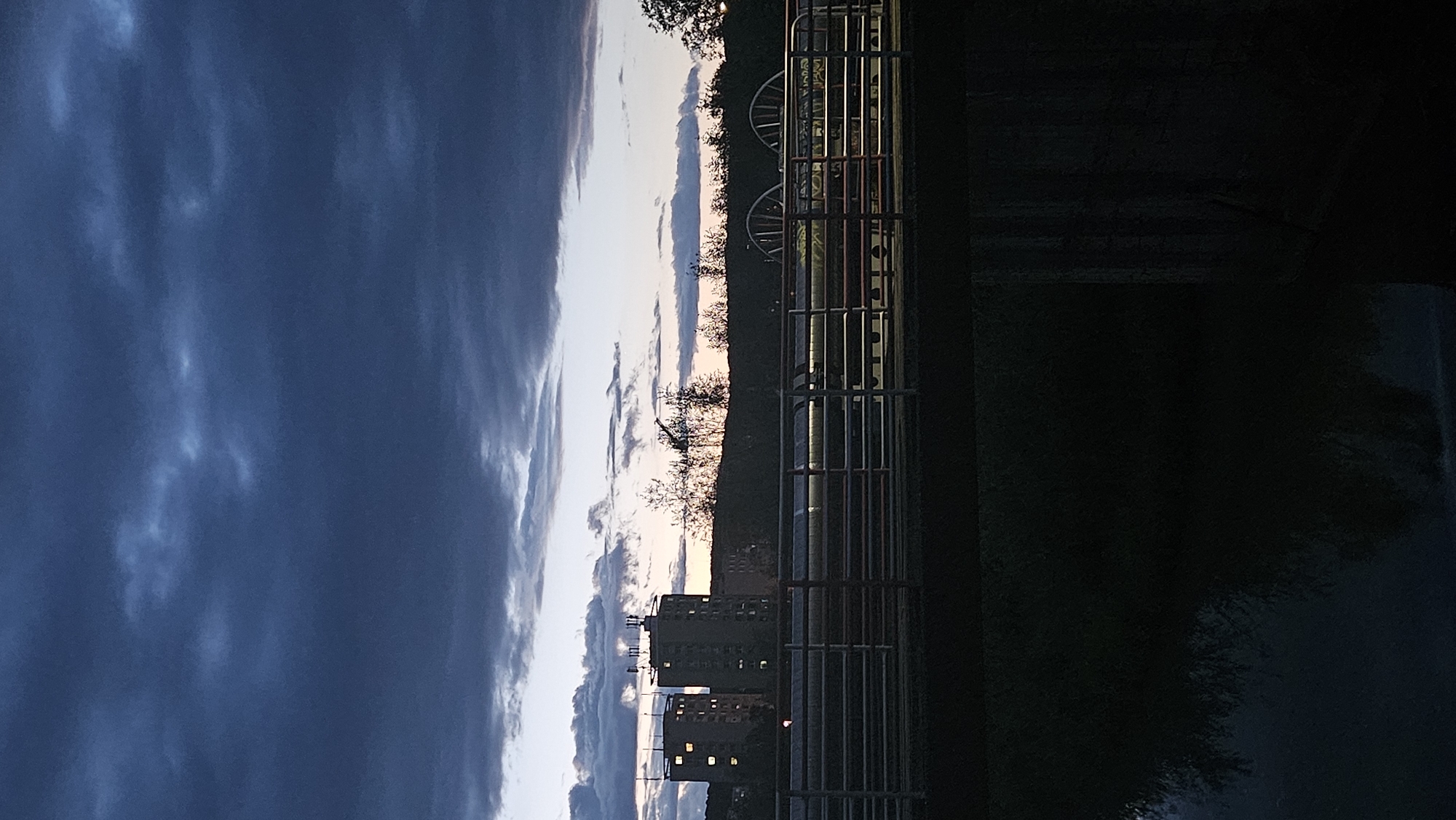The image depicts an exterior view of a body of water in the city of Prešov, Slovakia, during dusk. In the background, two tall, apartment buildings are visible, with lights illuminated in some windows. A row of trees forms a silhouette against the darkening sky, which is filled with dramatic, dark clouds. In the foreground, a bridge railing made of metal bars spans across the frame, with a darker structure below it. The water in the foreground is dark and reflects some of the scene above. Two large, circular structures, possibly industrial equipment or decorative elements, are partially visible behind the bridge railing. No people or animals are discernible in the image.