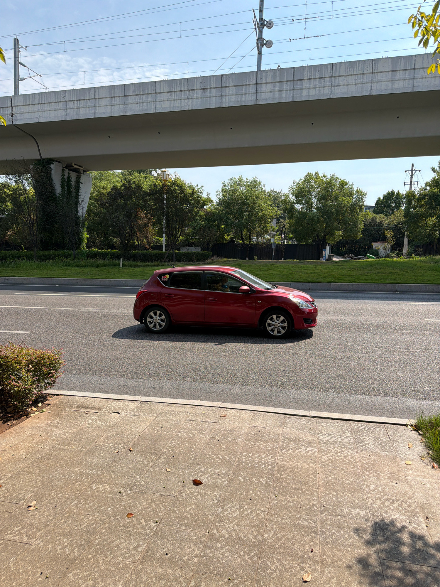 A red Nissan hatchback is driving on a paved road in Jiangjin, China. The car is a hatchback with four doors and appears to be in motion, as indicated by the slight blur of the wheels. It has a red paint job and silver alloy wheels. A red bull sticker is visible on the rear passenger window. The car is driving in the right lane of a multi-lane road. The road has white lane markings.
In the background, there is a large, elevated concrete structure, possibly an overpass or a bridge, with train tracks running along its top. This structure is supported by concrete pillars, one of which is partially covered in ivy. Beyond the overpass, there are trees and green foliage, suggesting a park or natural area. The sky is blue with some scattered clouds. The ground in the foreground is a paved walkway made of large square tiles, with fallen leaves scattered on it. The overall scene depicts an urban or suburban environment during the daytime.