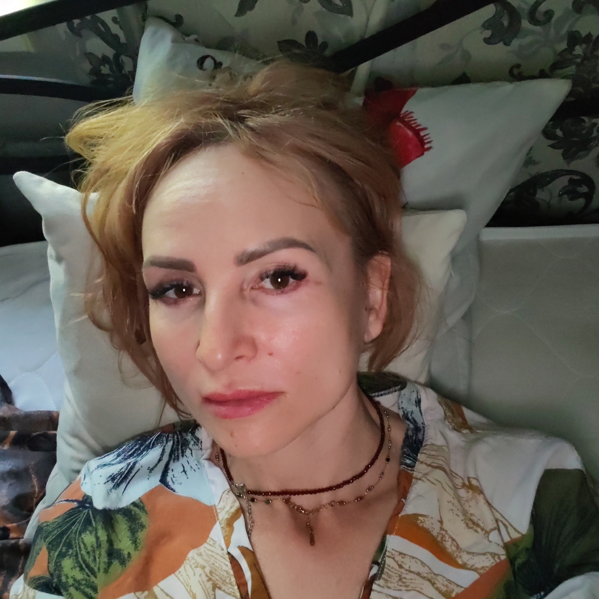 A woman is lying on a bed. She has fair skin, reddish-blonde hair pulled up into a messy bun, and is wearing makeup. She is wearing a floral-patterned top and two necklaces. The bed has white pillows and a patterned headboard. The photo was taken indoors. There is no evidence to suggest that the photo was taken in Chaikovsky, Russia.