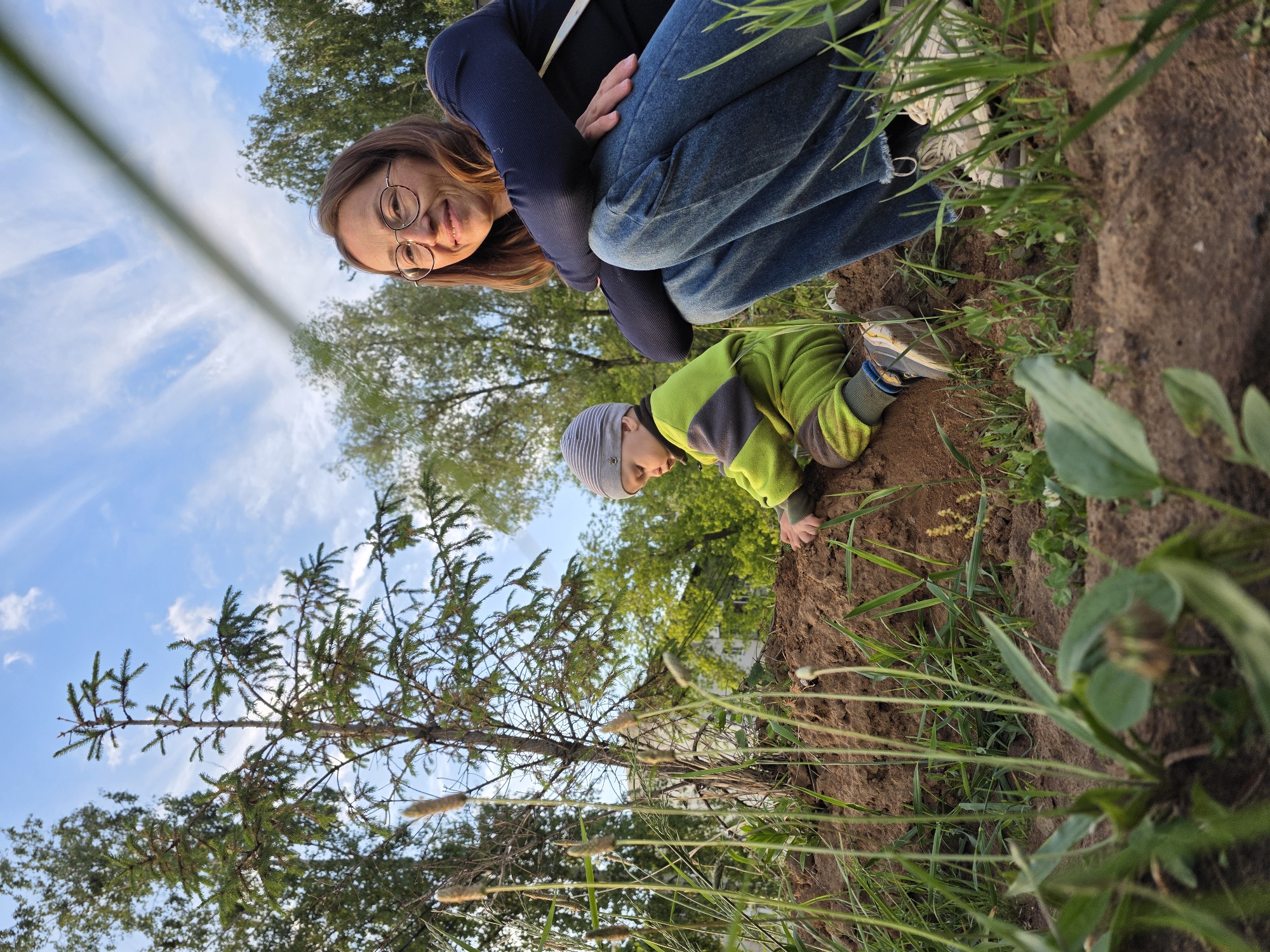 The image depicts an adult and a child outdoors in a natural setting. The adult, wearing glasses, a dark long-sleeved shirt, and jeans, is crouched down near the edge of a dirt area. The child, wearing a light green and gray jacket and a striped hat, is kneeling and appears to be touching the dirt. There is grass and other foliage present. Trees are visible in the background against a partly cloudy sky. The location is Chaikovsky, Russia.