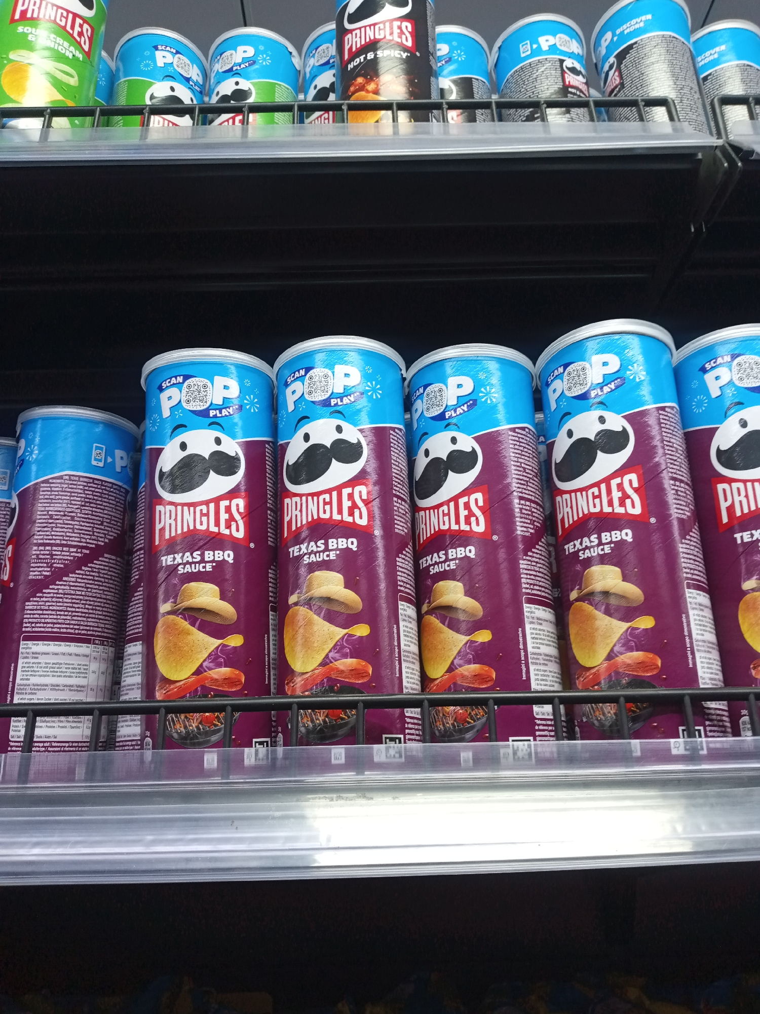 A retail shelving unit, located in Kawaji, Nigeria, displays multiple cylindrical snack food containers. The top shelf contains a row of Pringles brand potato crisp cans. From left to right, a green can labeled "SOUR CREAM & ONION" is visible, followed by several blue cans with "SCAN POP PLAY" branding and Pringles' mascot Mr. P's stylized face. Further right, a red and black can is labeled "HOT & SPICY," adjacent to more blue Pringles cans, some partially obscured. The top shelf features a metal support bar.
The bottom shelf presents a distinct row of Pringles "TEXAS BBQ SAUCE" flavor cans. These cans are predominantly purple with a light blue top featuring "SCAN POP PLAY" and a QR code. Each purple can displays a graphic of Mr. P wearing a cowboy hat, a golden potato crisp, and a depiction of grilled meat on a barbecue grate. The white Pringles logo is prominent below Mr. P. The back of the cans reveals nutritional information and product details printed in various languages. The bottom shelf also features a clear plastic price strip holder and a metal support bar. The lighting appears uniform and artificial. No people are visible in the scene.