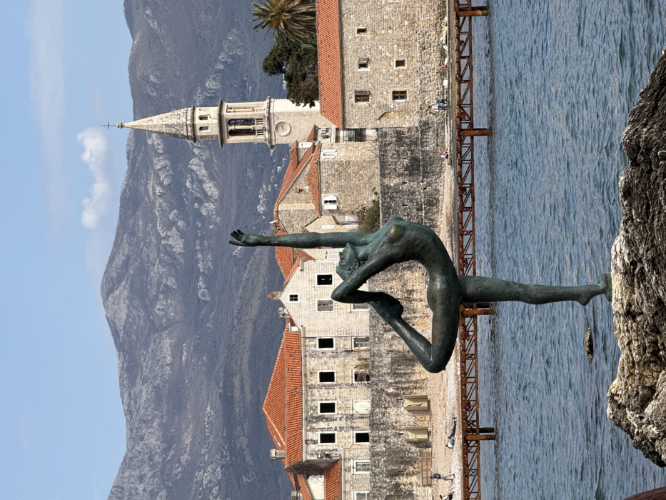 The image displays the "Dancer from Budva" bronze statue positioned on a rock outcrop in the Adriatic Sea, in front of Budva's Old Town in Montenegro. The statue, exhibiting a greenish patina, depicts a slender female figure balancing on her left leg, with her right leg raised backward and upward, and both arms extended vertically overhead.
Behind the statue, the fortified stone walls of Budva's Old Town rise directly from the sea. Numerous stone buildings with pitched terracotta roofs are visible within the Old Town. A prominent light-colored bell tower with a pointed spire and visible clock faces, identified as part of St. Ivan Church, stands on the right side of the Old Town. A narrow, pebbled beach runs along the base of the Old Town walls, adjacent to a weathered wooden pier or walkway supported by multiple pilings extending into the water.
Several small, indistinct human figures are present on the beach and near the pier; some are standing, while others appear to be lounging on the sand. The sea in the foreground and mid-ground is choppy and blue. The background consists of a range of large, rugged grey mountains with sparse vegetation, under a clear, light blue sky with minimal cloud cover. The scene is illuminated by natural daylight.
