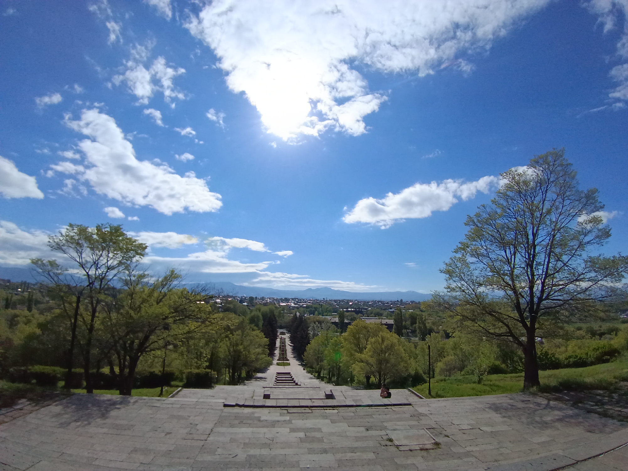 The image shows a wide-angle view of the Cascade in Yerevan, Armenia. The foreground features stone steps leading down the structure. On either side, there are trees with green foliage. In the background, the city of Yerevan stretches toward the horizon, with mountains visible in the distance under a blue sky dotted with clouds and bright sunlight. There's a person sitting on the steps.