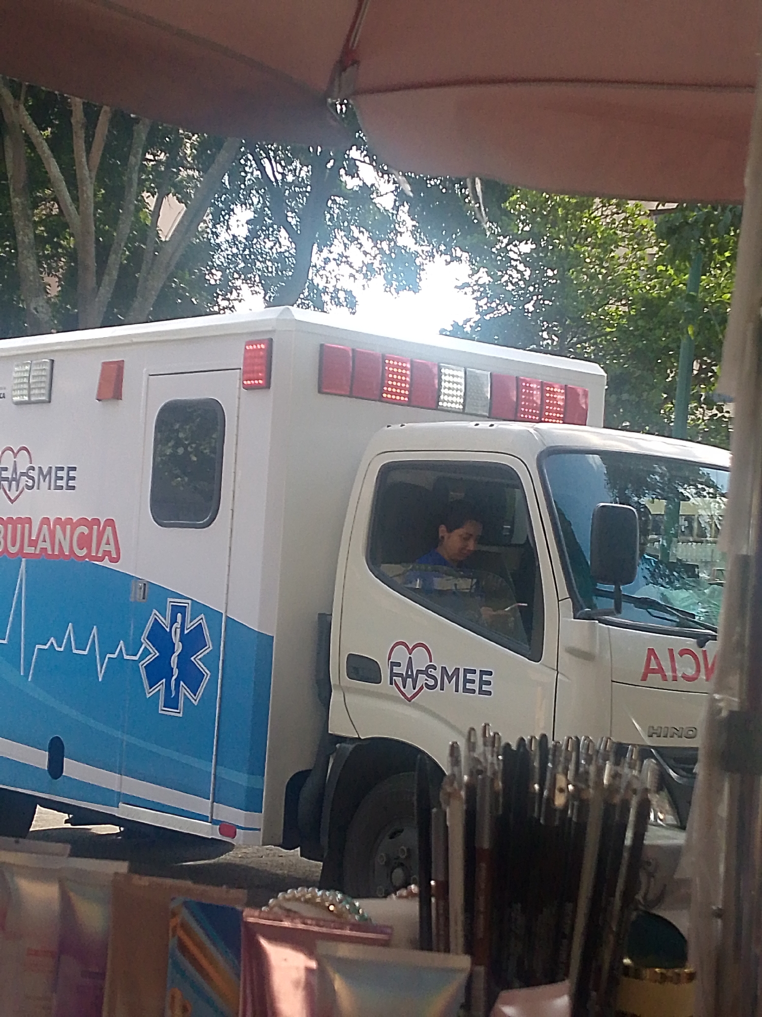 The image captures a side view of a white ambulance, likely in an urban setting, given the presence of trees and what appears to be a street or paved area. The ambulance has the word "AMBULANCIA" written in large red letters on its side, along with a logo featuring a heart and the text "EASMEE". A prominent blue Star of Life symbol is also visible on the rear section of the ambulance, below a heartbeat EKG line graphic.
Inside the driver's side of the cab, a person wearing a blue shirt is seated, looking down at something in their hands. They appear to be wearing earbuds. The ambulance has red and white flashing lights mounted on its roof.
In the foreground, partially obscuring the lower part of the ambulance, is a collection of makeup brushes arranged in a container. The background shows lush green trees with dappled sunlight filtering through. The overall impression is that of a street scene in a city, potentially during daylight hours, with the ambulance being the primary subject. While the provided metadata indicates the location as Caracas, Venezuela, this cannot be definitively confirmed from the visual elements within the image alone.
