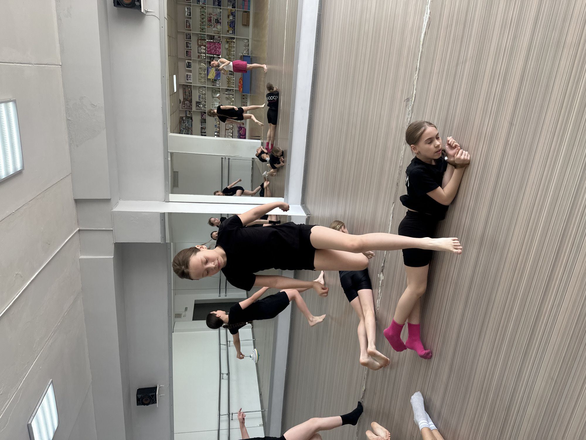 The image depicts a dance studio in what appears to be Votkinsk, Russia. Several young girls dressed in black dance attire are present, with some performing dance moves and others resting. A large mirror covers one wall, reflecting the dancers and the surrounding environment, including shelving units filled with memorabilia. The wall adjacent to the mirror has a wood-like paneling. One girl is wearing bright pink socks and another is wearing white socks. The lighting in the studio appears to be provided by overhead fluorescent lights.