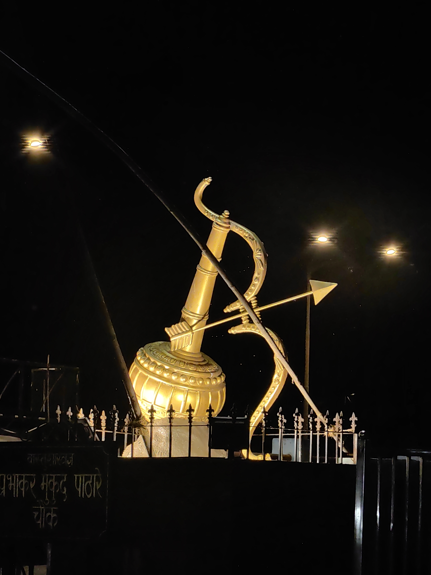 The image depicts a golden statue of a bow and arrow, illuminated at night. The statue is located at Prabhakar Mukund Pathare Chowk in Mumbai, India. The statue is positioned behind a black fence with decorative elements. In the background, there are several lights, including two prominent bright lights in the upper right quadrant of the image, and a diffuse light source in the upper left quadrant. The sky is dark, indicating nighttime. Below the fence, a black sign with white text is visible, which appears to be a name or inscription in Marathi. The overall scene is stark and illuminated, highlighting the golden statue against the dark night.
