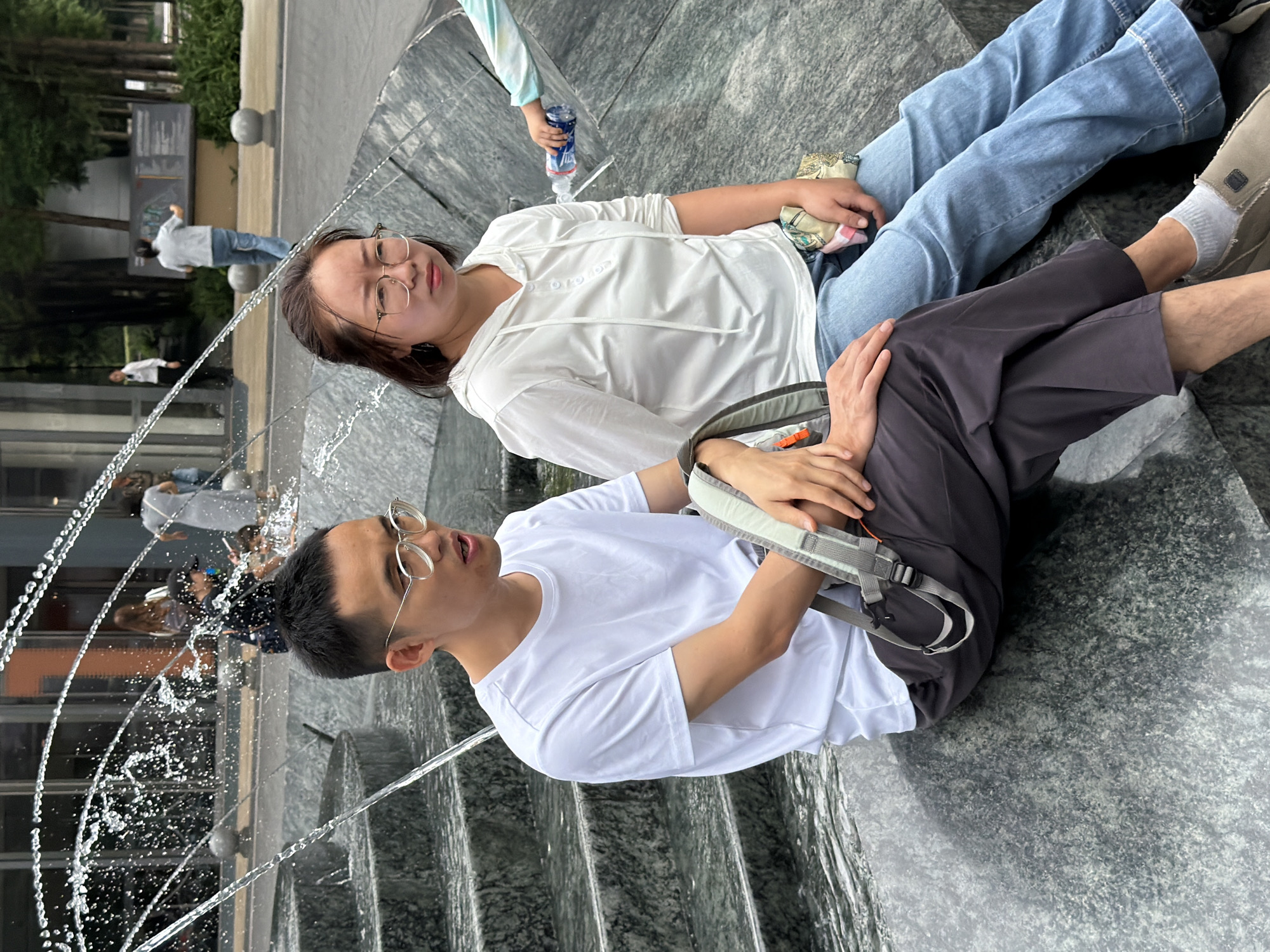 In Yuzhong District, China, a man and a woman are seated on a tiered stone structure next to a fountain with water jets. The man, wearing glasses, a white t-shirt, and dark pants, is on the left. He has a grey backpack with straps visible. The woman, also wearing glasses and a white hooded shirt, sits to his right, dressed in blue jeans. Her hands are resting on her lap, and she holds a small, patterned cloth object. Behind them, other people are visible in the background, including a woman in a grey dress and a man in a white shirt. The environment appears to be an outdoor public space, possibly a plaza or park, with buildings and trees in the distance. The fountain is actively spraying water.