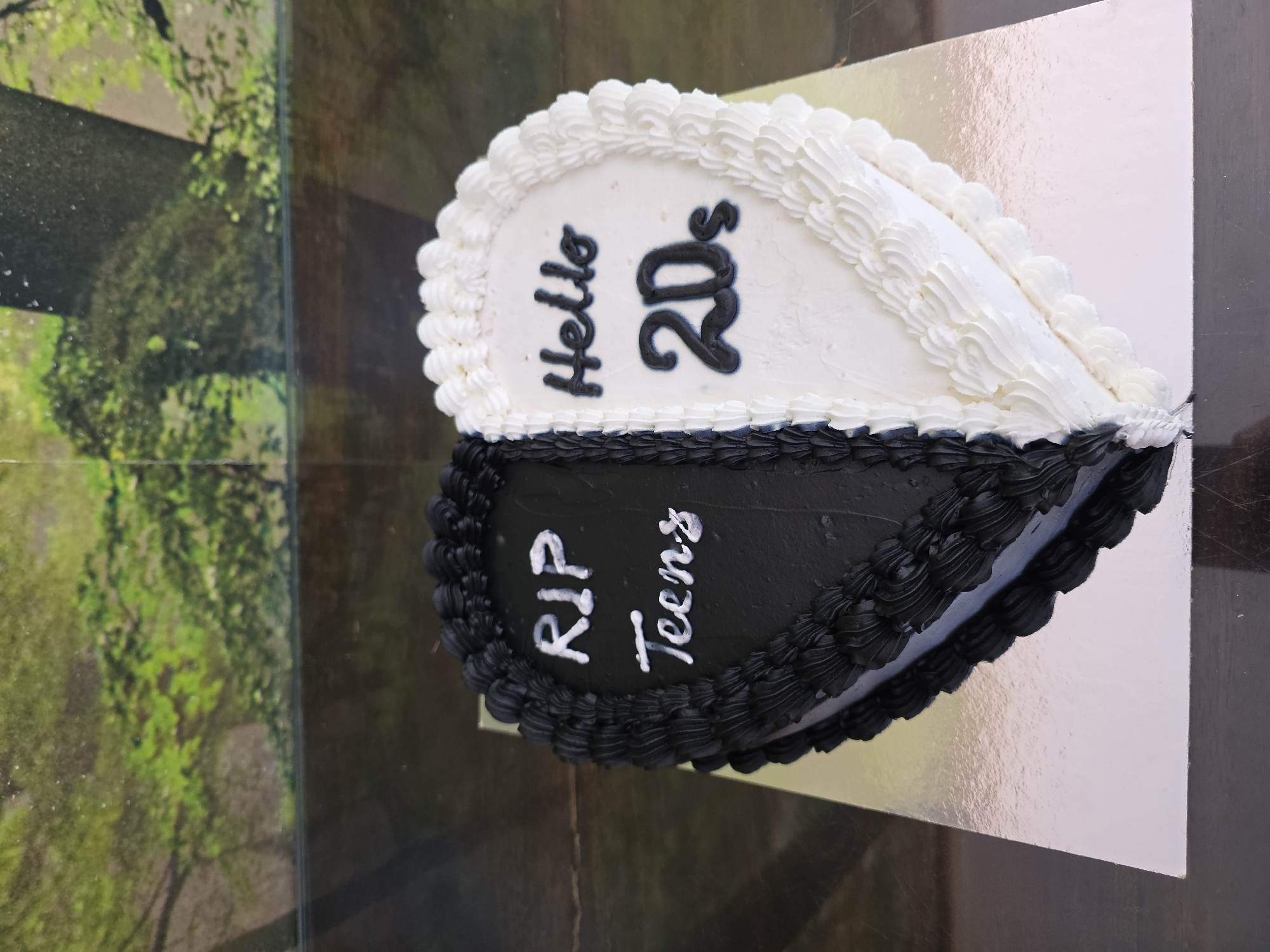 A heart-shaped cake sits on a silver-colored cardboard base on a reflective surface. The cake is divided into two halves: the upper half is white, with "Hello 20s" written in dark frosting, and the lower half is black, with "RIP Teens" written in white frosting. Both halves are decorated with elaborate frosting around the edges. The reflective surface reflects a blurred image of trees and foliage. The photograph was taken indoors, and the exact location in Varna, Bulgaria, cannot be determined from the image.
