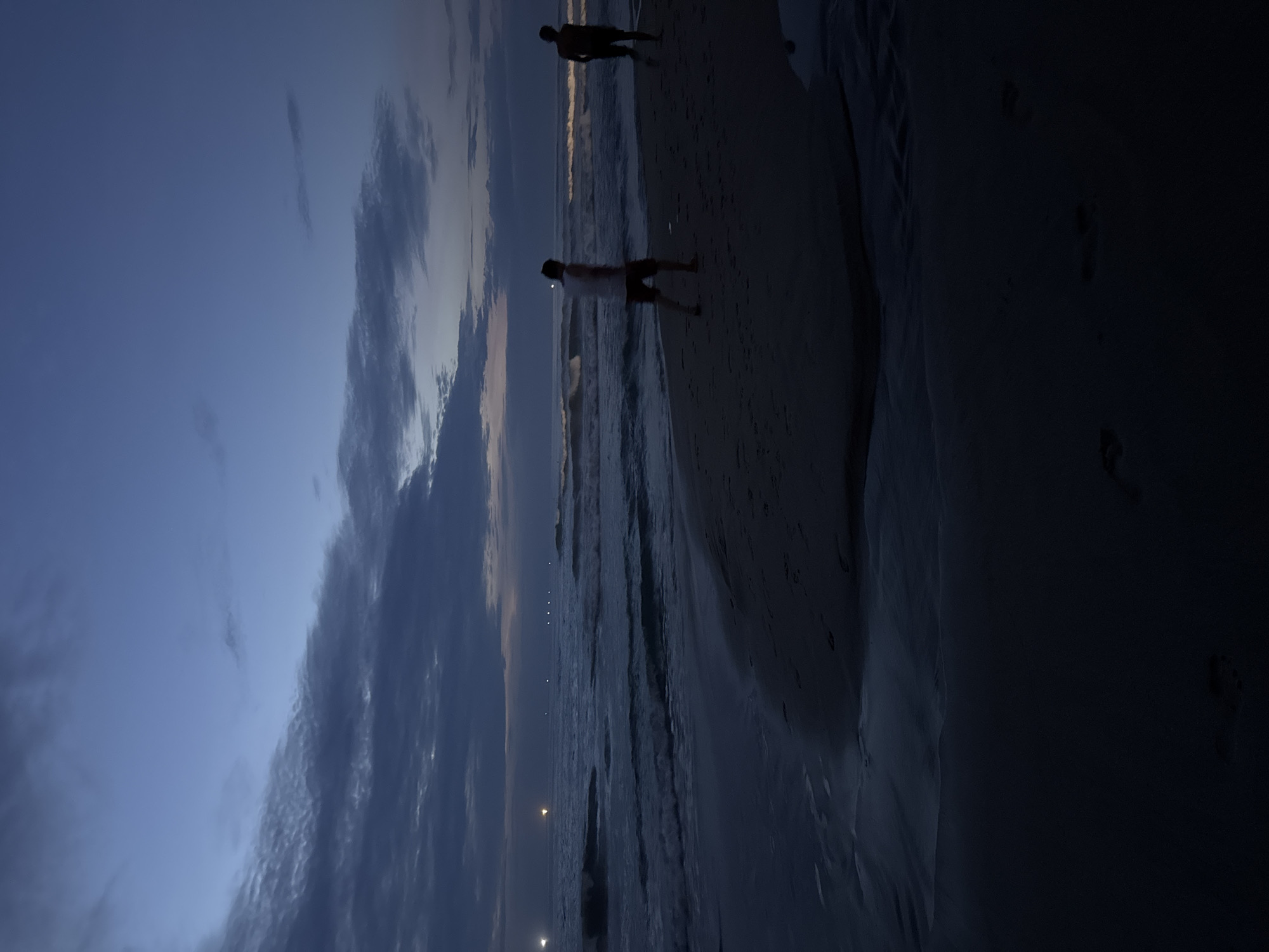 The image depicts a beach at dusk or night. Two figures are silhouetted against the receding waves. The sky above is a mix of dark blue and clouds. There are lights visible in the distance over the water. Footprints are visible in the sand on the right side of the frame. The location appears to be coastal but the city of Da Nang cannot be verifiably confirmed.