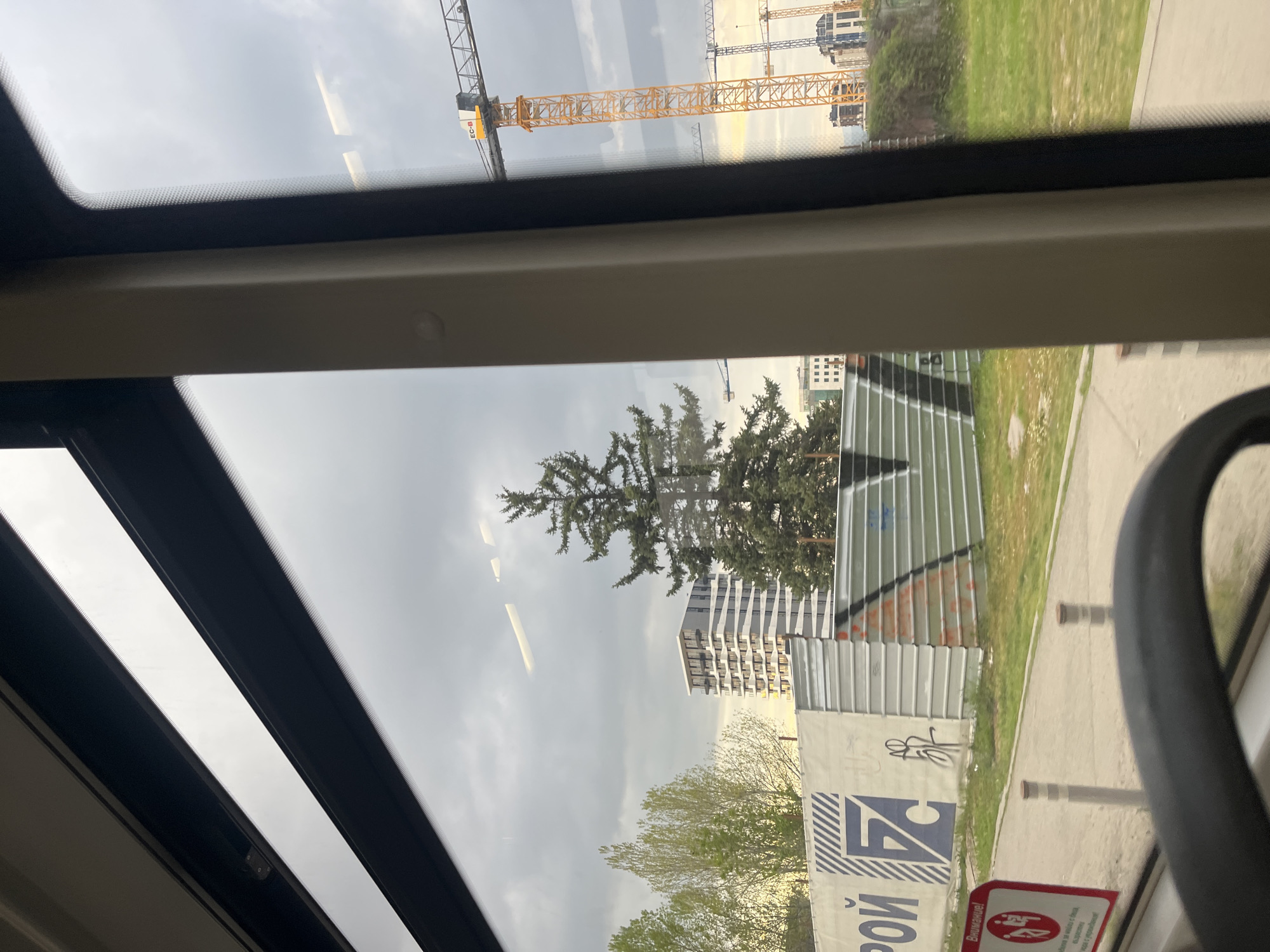 This media file presents a view from inside a vehicle, likely a bus or tram, looking out through a large window at an urban landscape in Sofia, Bulgaria. The image, initially rotated counter-clockwise, shows a scene dominated by construction and development under an overcast sky.
In the foreground, parts of the vehicle's dark window frame are visible on the top and bottom edges, with a dark, curved handle or bar on the right side. Reflections on the window subtly hint at the vehicle's interior or the sky above.
The midground features a large, dark green pine tree on the left. To its right, several modern multi-story residential buildings are visible, characterized by their angular designs and visible balconies, suggesting recent construction. Below these buildings, a corrugated metal construction fence runs along a grassy verge and a paved sidewalk, where short, grey bollards are positioned.
The background is filled with signs of ongoing development: numerous large, yellow tower cranes pierce the grey, cloudy sky. One crane boom prominently displays the text "EC-B". The overcast weather suggests a cloudy day, making it difficult to determine the exact time of day, though it is clearly daylight.
Visible text on a large white banner attached to the construction fence includes Cyrillic script: "СТРОЙ" (STROJ, meaning "BUILD" or "CONSTRUCTION") and a distinct blue logo resembling "EZ" or "FZ" with diagonal stripes. Below this, a red warning sign reads "Внимание!" (Vnimanie!, "Attention!" or "Warning!"), followed by "Опасно за здравето и живота." (Opasno za zdraeto i zhivota., "Dangerous for health and life."), accompanied by a pictogram of a falling person, emphasizing safety hazards at the construction site. The overall scene captures a moment of urban expansion and development in Sofia.