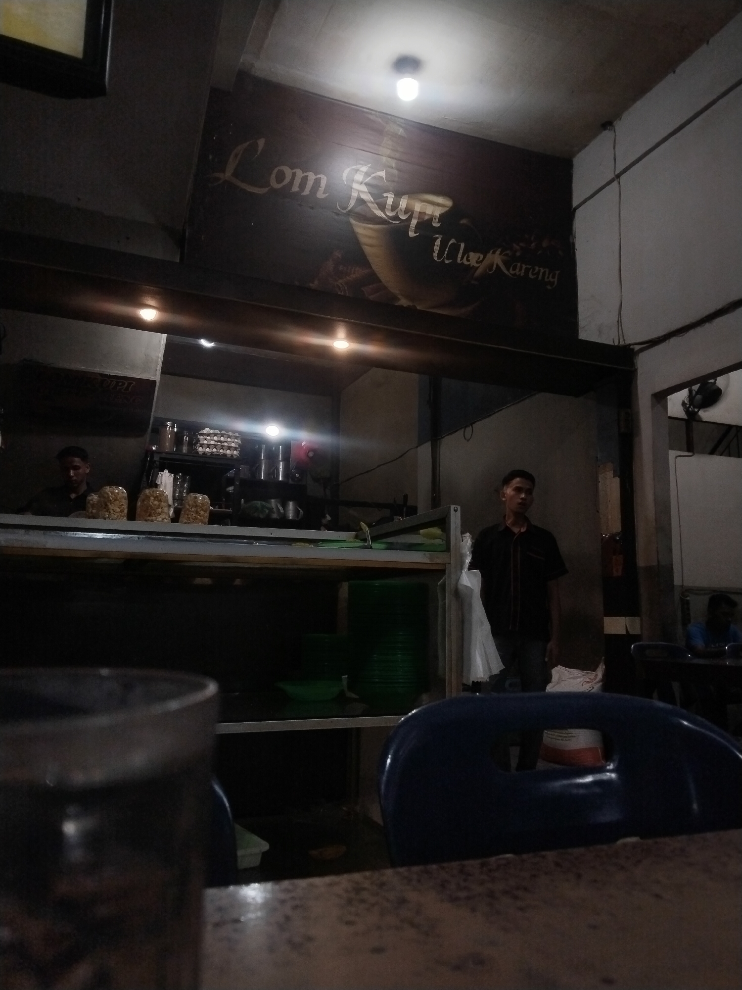 The image shows the interior of "Lom Kupi Ulee Kareng", a cafe or restaurant located in Lhokseumawe, Indonesia. A sign with the establishment's name is visible above a service counter. Two men are present; one behind the counter, and another standing near the entrance to a dining area. The interior features simple furniture including blue plastic chairs. A drinking glass is placed on a table in the foreground. The scene appears dimly lit.