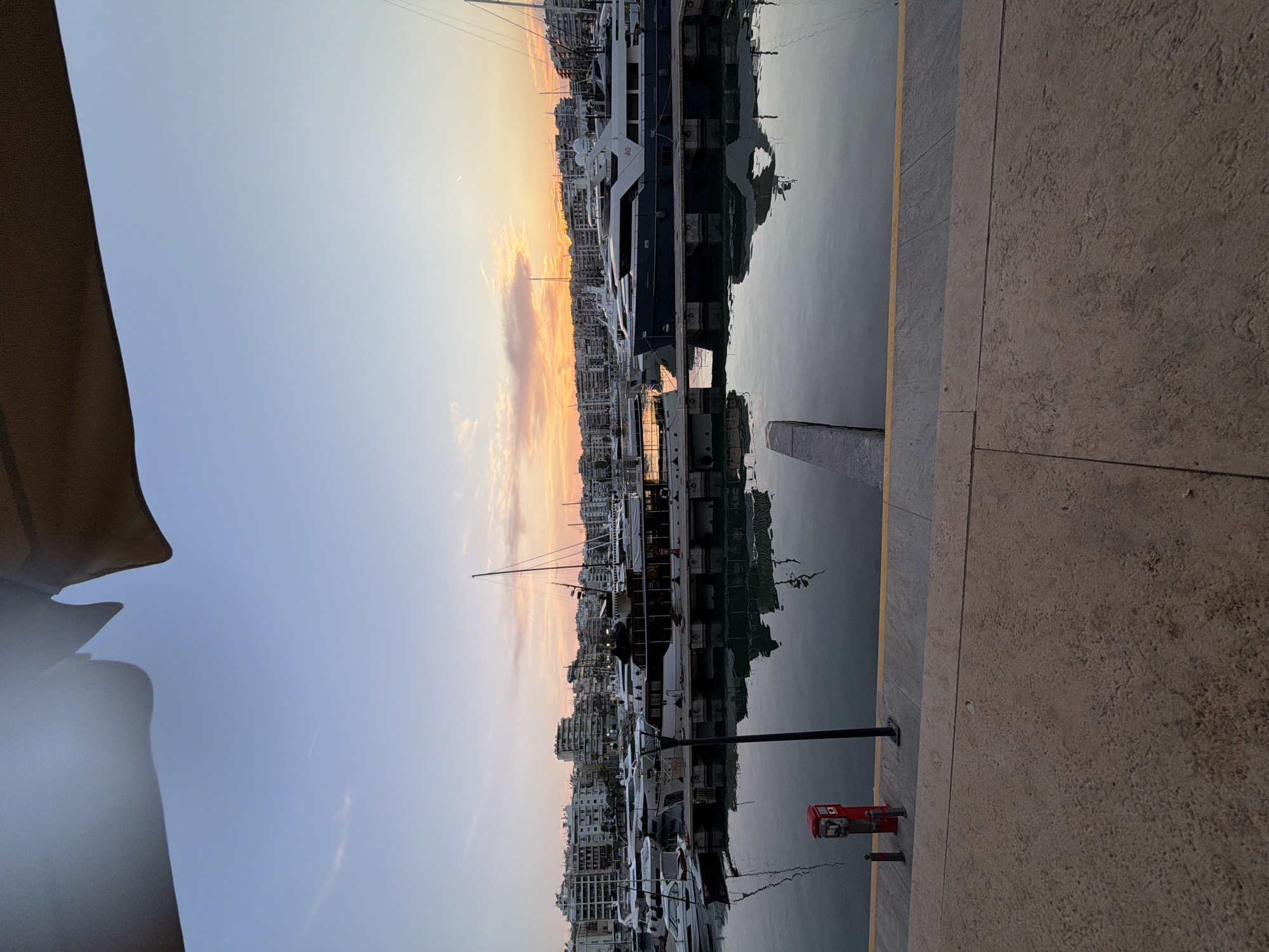 The image shows a view of a marina in Piraeus, Greece, at sunset. The sky is a gradient of blue to orange with clouds present. Multiple yachts and boats are docked along the waterfront, with buildings visible behind them. The water reflects the boats and the sky. The foreground consists of a concrete walkway or dock. A red utility box is visible on the walkway. A portion of a tent or shade structure is visible in the upper left. The photo is taken from a ground-level perspective.