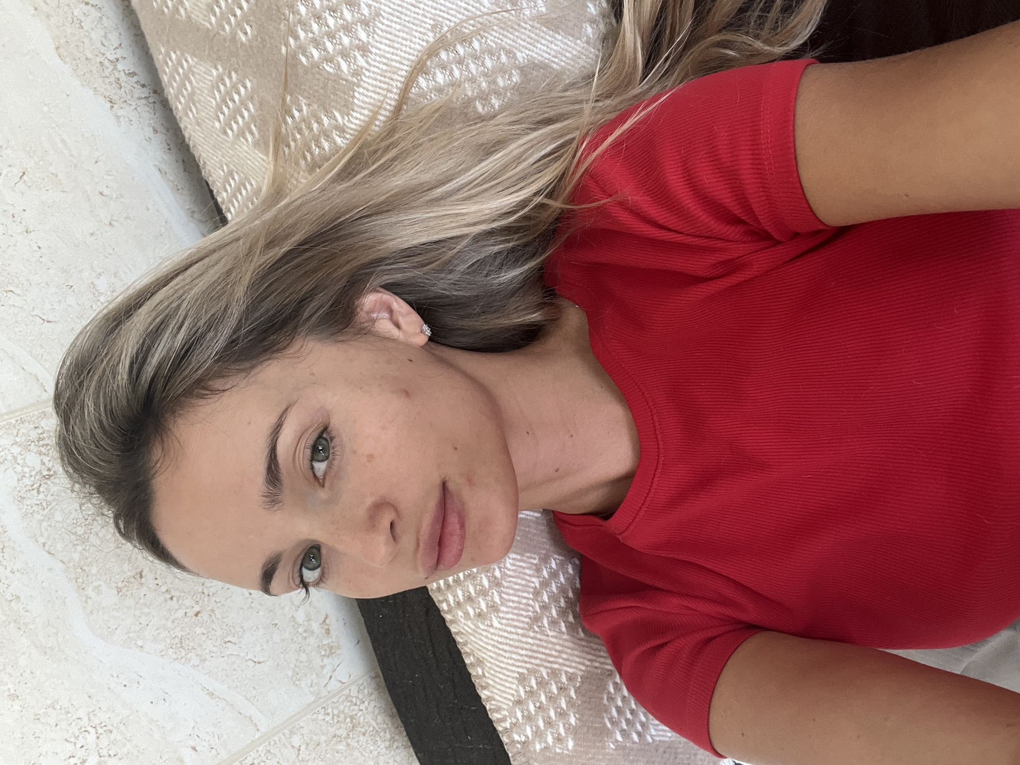 A young woman with long, blonde hair and green eyes, wearing a red t-shirt, lies on her back on a bed. She is looking directly at the camera with a neutral expression. The room appears to be a bedroom, with a tiled wall and a textured blanket in the background. There are no other people or objects in the image.
