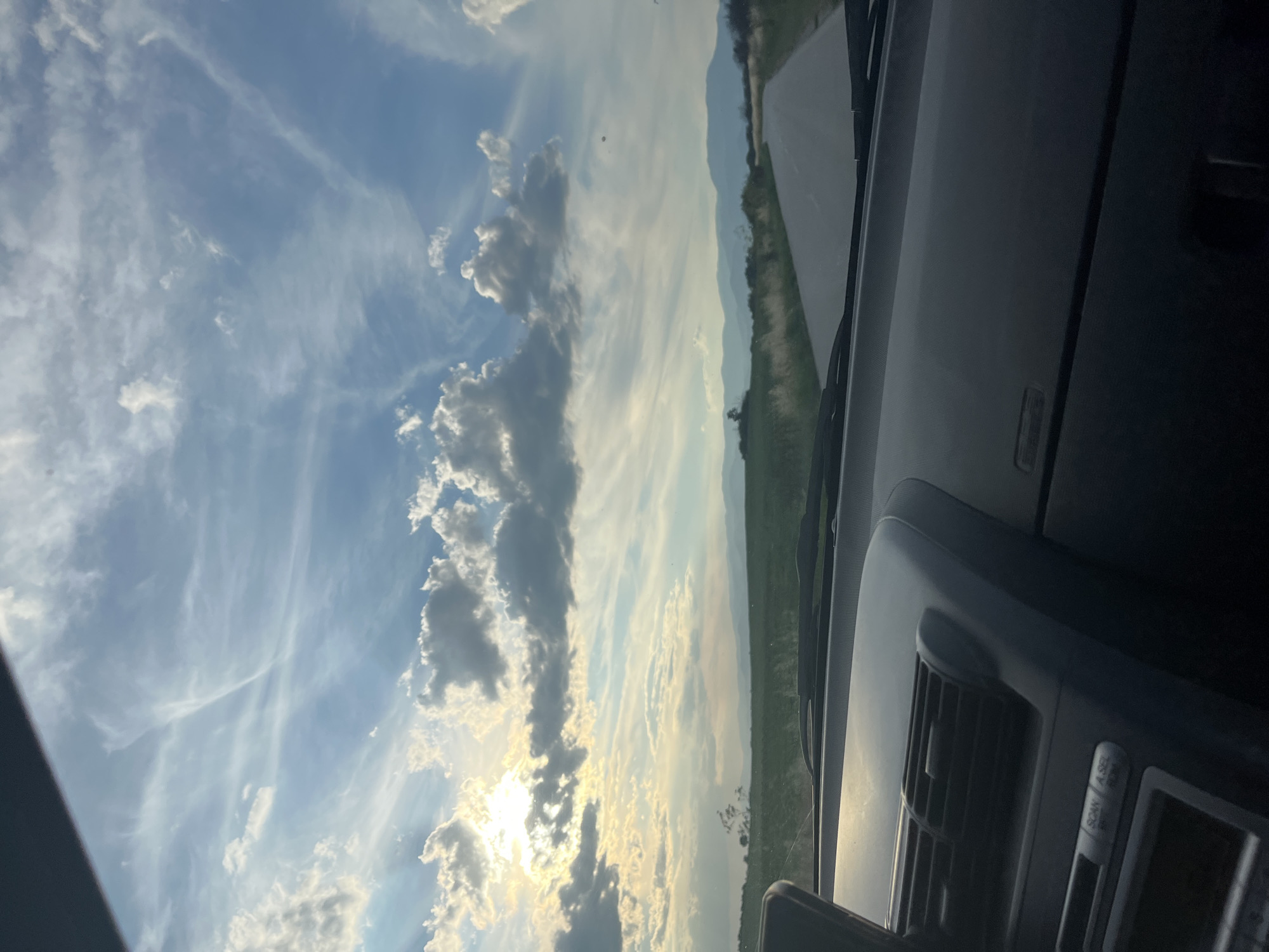 A wide-angle, low-angle perspective from the interior of a vehicle shows a road stretching towards distant mountains under a sky filled with dramatic clouds. The sun is partially obscured by clouds on the left side of the frame, casting a warm glow over a green, hilly landscape. The road appears to be asphalt and curves slightly to the right. The dashboard of the vehicle is visible in the foreground, with a navigation device mounted on the left and an air vent. The location is identified as Zhitnitsa, Bulgaria. No people are visible in the image.