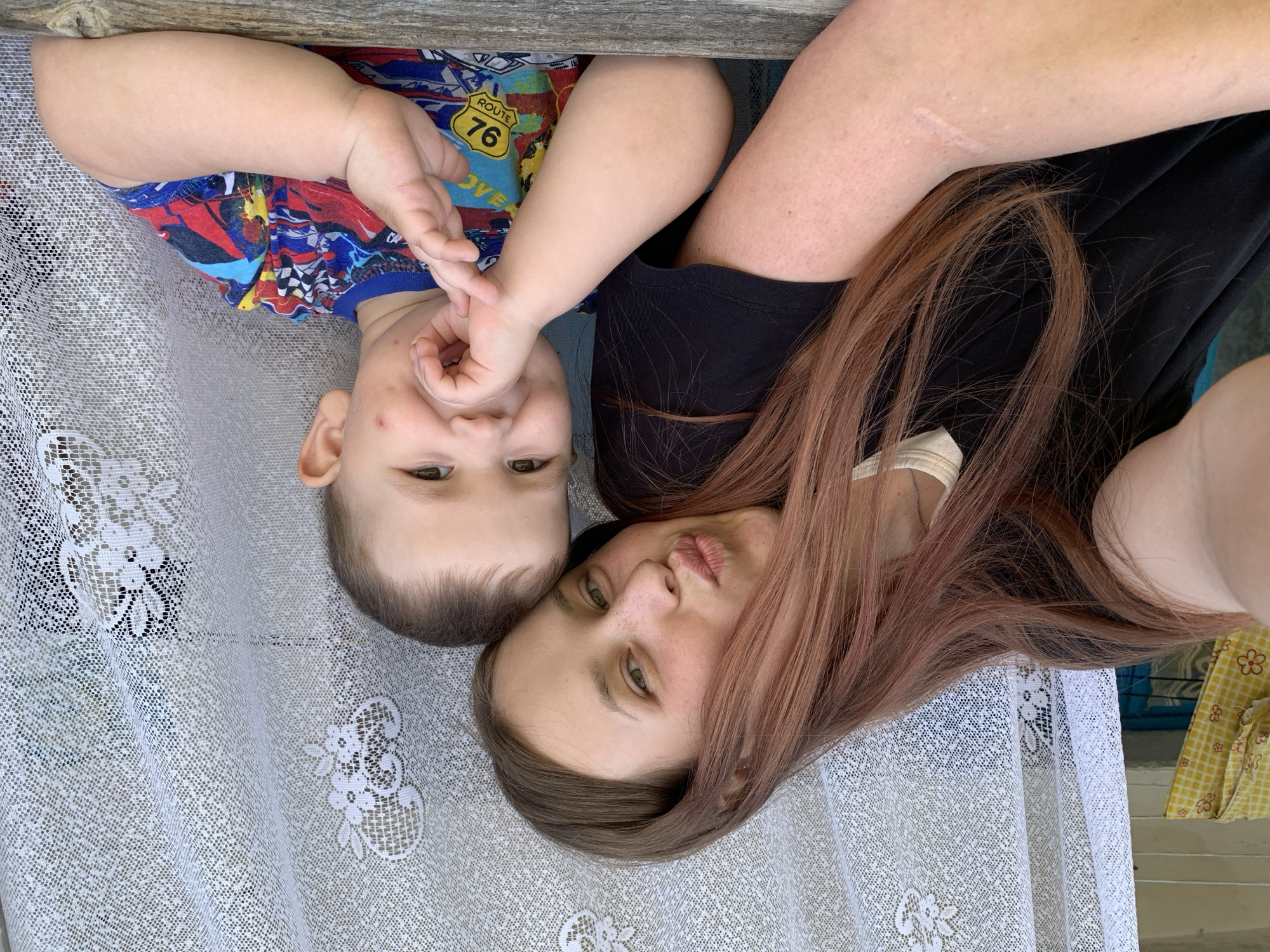 A woman and a young child lie side-by-side on a white lace-patterned surface, possibly a tablecloth or blanket. The woman, on the right, has long hair with brown roots and reddish-brown highlights. She is wearing a black top and has fair skin. The child, on the left, has a fair complexion and short brown hair. He is wearing a colorful t-shirt with images of cars and the number "76". The child's hand is raised near his face. The background is blurred. The location and city of Фоки, Russia cannot be determined based on the image.