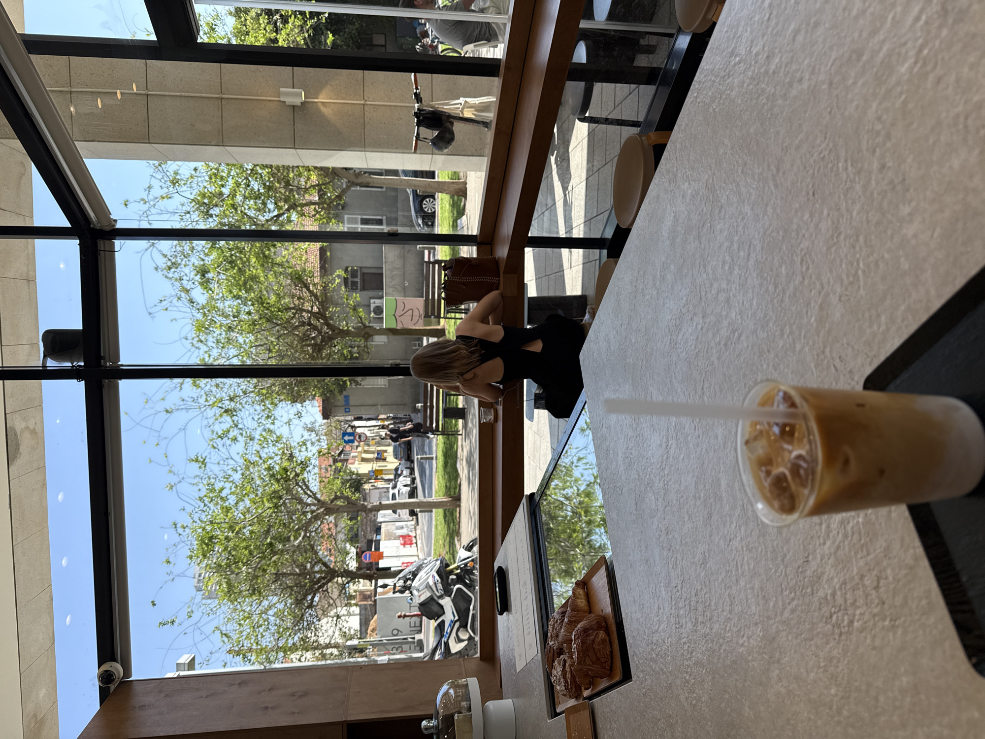 The image captures a scene from inside a modern cafe in Tel-Aviv, Israel, looking out through expansive floor-to-ceiling windows onto an urban street.
In the foreground, on the right, a textured light-colored countertop extends into the frame. An iced coffee with a straw is prominently placed on a dark tray, next to a wooden board laden with an assortment of pastries, including several croissants. A display screen on the counter reflects elements of the exterior.
A woman with long, light-colored hair, dressed in a black top, sits at the counter with her back mostly to the camera, looking out the large windows. She appears to be enjoying a moment of relaxation.
Beyond the windows, a bustling but peaceful street scene unfolds under a bright, clear sky. Green trees with full foliage line the street, and various vehicles, including cars and a motorcycle, are visible. Buildings with architectural details, some showing air conditioning units, stand across the street. Visible street signs include blue directional arrows and a red stop sign. Faint text "NET" and the number "39" are discernible on a building facade in the distance. A security camera is mounted on the interior window frame.
The bright daylight indicates it is likely morning or afternoon. The overall atmosphere is calm and inviting, characteristic of a cafe in an urban setting.