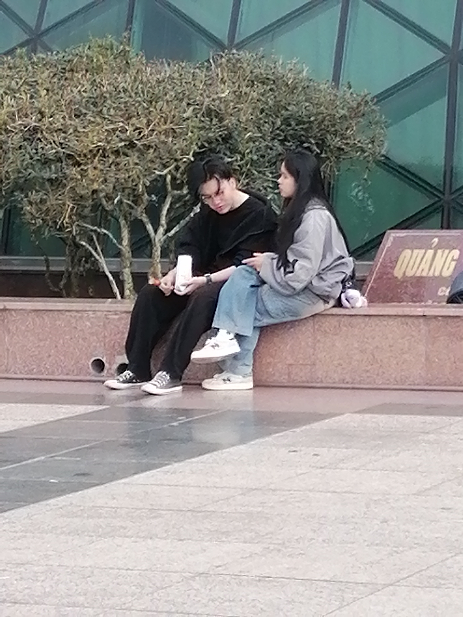 This is a medium shot, eye-level photograph of two young adults sitting on a low, reddish-brown concrete wall outdoors. The background consists of a large, dark green building with a geometric pattern and a dense, dry-looking shrub behind the wall.
The person on the left appears to be a male, wearing a black long-sleeved shirt and black pants. He has dark hair and is wearing glasses. He is holding a white object, possibly a phone or a small box, in his left hand and his right hand is resting on his lap.
The person on the right appears to be a female, wearing a light grey jacket over a darker top and light blue jeans. She has long, dark hair. She is looking to her left, away from the camera.
Both individuals are wearing sneakers. The male is wearing black and white high-top sneakers, and the female is wearing white sneakers.
To the right of the subjects, a rectangular sign is partially visible. The sign is reddish-brown with yellow lettering that reads "QUANG". Below that, there is a smaller line of text in white.
The ground in front of the wall is paved with large, square tiles, with some appearing to be wet.
The overall impression is a casual, outdoor scene with two people sharing a moment. The specific location is not explicitly identifiable from the image, though it is stated to be in Da Lat, Vietnam.