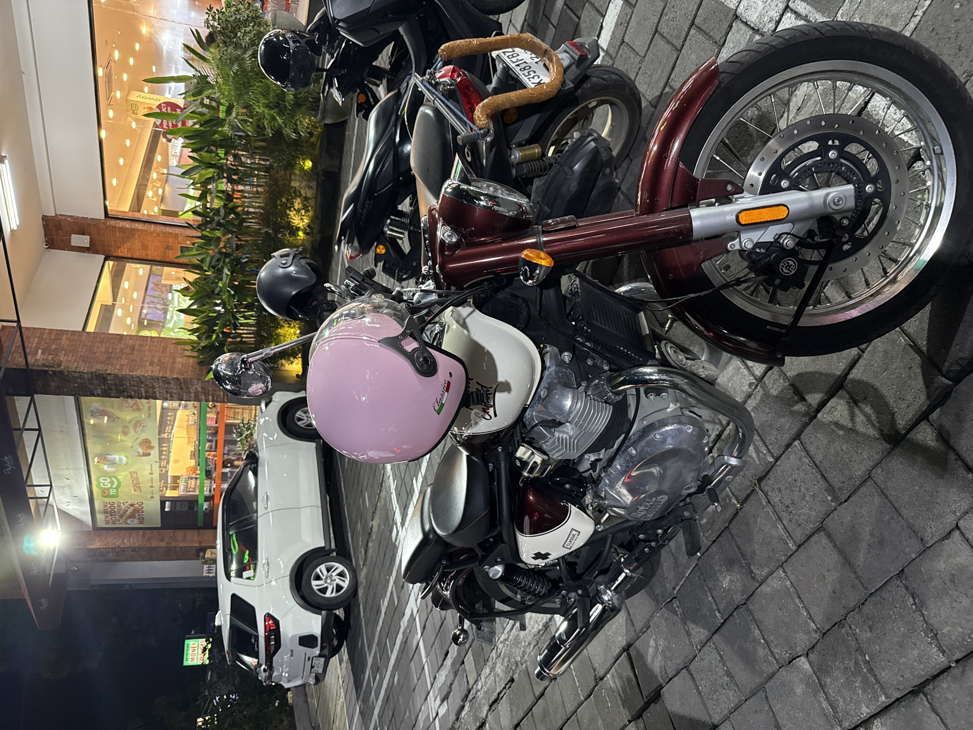 The image captures a night scene featuring several parked motorcycles and a car outside a commercial establishment. In the foreground, a dark red/maroon custom-style motorcycle, possibly a cruiser, is prominently displayed. Two helmets rest on its seat area: one pink with "Classica" written on it, and a white one with "Classic" visible on its side panel. The engine casing of this motorcycle clearly shows the text "ROYAL ENFIELD". Behind it, part of a black scooter is visible, sporting a license plate that reads "K3581FBZ". The handle of this scooter appears to be wrapped in a brown material.
To the left, a white SUV or car is parked, reflecting the bright lights from the adjacent buildings. The ground where the vehicles are parked is paved with dark grey interlocking bricks.
The background consists of brightly lit storefronts with large glass windows, suggesting a fast-food restaurant or a convenience store. Green plants in planters line the front of the buildings. Visible text on the storefronts and signs includes "NEW MENU HOTDOG", "GO TO", "HOTDOGS", and "MONEY" on a green sign. Another sign on a building to the far left partially displays "Papade" and "Your way.". The overall lighting indicates it is nighttime, with artificial lights illuminating the scene and reflecting off surfaces. No people are visible. The location context is unknown.