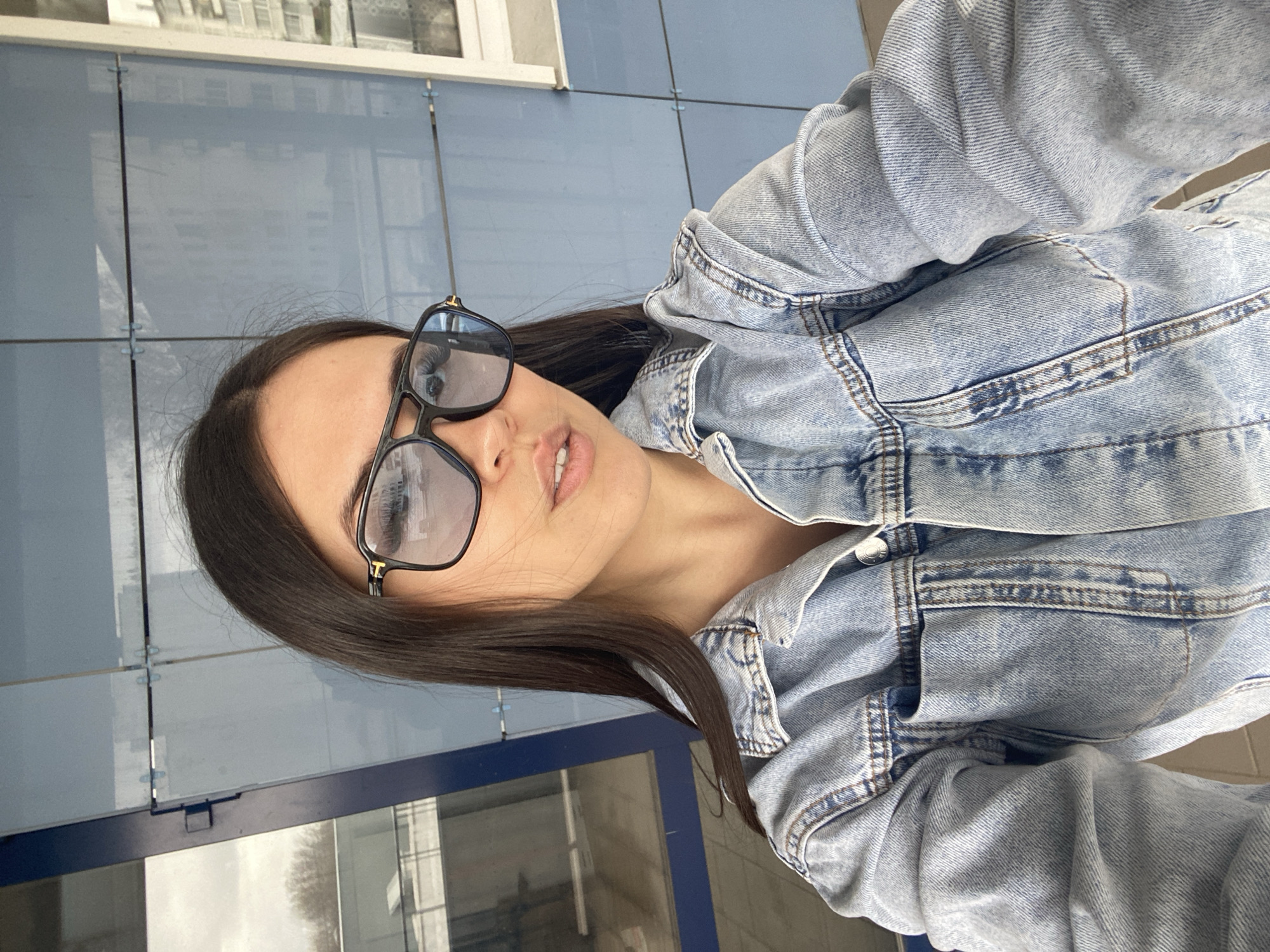 A woman with long, dark hair and sunglasses poses for a selfie. She wears a denim jacket. The background shows a building with blue tile panels and windows. The location appears to be in Chaikovsky, Russia, based on potential reflections in the glass.