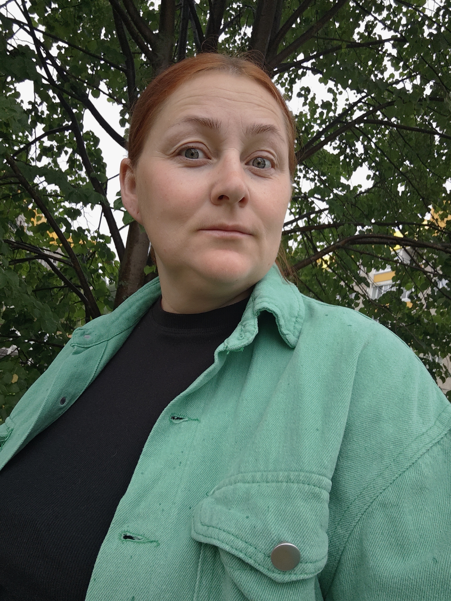 A medium shot shows a woman with red hair and light-colored eyes looking at the camera. She is wearing a black shirt and a teal jacket. The background is out of focus, showing green foliage and a building. The image was taken outdoors in what appears to be an urban setting.