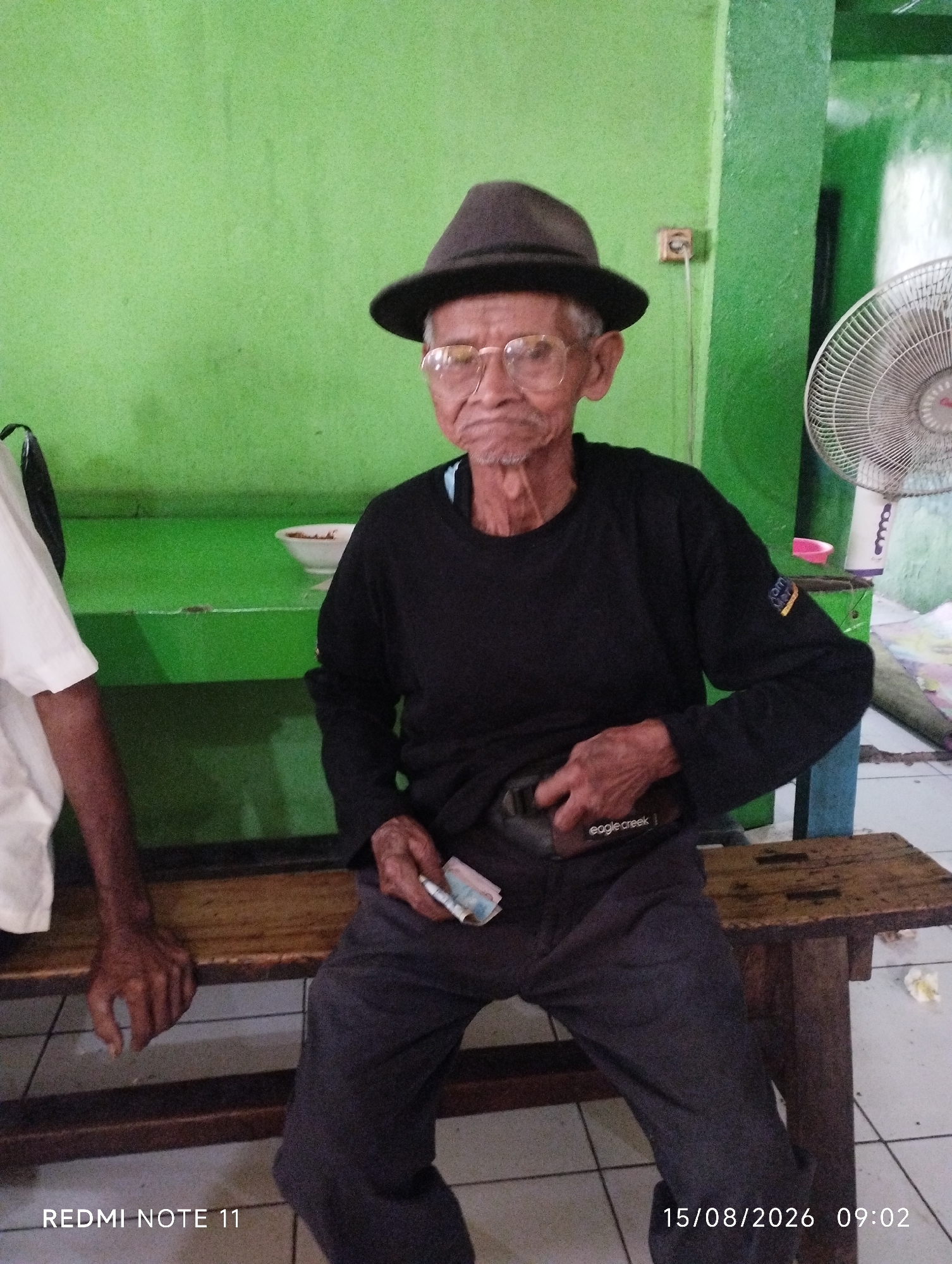An elderly man wearing a dark hat and glasses, and a black long-sleeved shirt, is seated on a wooden bench. He is holding a small stack of bills in his right hand and reaching into his pocket with his left hand, near a black pouch with "eagle creek" printed on it. The man's trousers are dark gray. To his left, another person, partially visible, wears a white shirt. The background consists of a bright green wall and a white fan. The floor is tiled. A wooden table is situated to the right of the man. The timestamp on the image reads "15/08/2026 09:02" and "REDMI NOTE 11". The scene appears to be indoors, possibly in a casual setting or community area. The image is consistent with being taken in Karawang, Indonesia, given the timestamp format and phone model, which are common in the region.