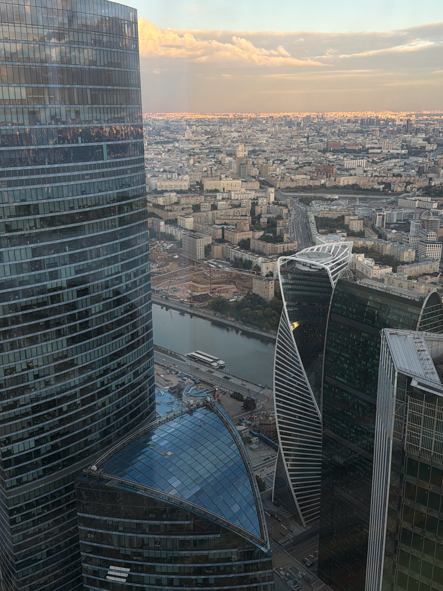 The image captures a panoramic view of the Moscow city skyline at dusk, likely from a high vantage point in the Moscow International Business Center (MIBC), also known as Moscow-City, located in the city of Khimki, Russia. The foreground is dominated by the glass facade of a modern skyscraper, its reflective surface mirroring the surrounding urban landscape and the pastel hues of the sky.
Several other contemporary architectural structures are visible, including a distinctive twisted skyscraper with white detailing and another building with a dark, faceted glass exterior. These buildings are situated near a body of water, possibly the Moskva River, which snakes through the urban expanse. A bridge and some boats are discernible on the water.
The midground showcases a dense collection of residential and commercial buildings, characteristic of a large metropolis. The cityscape stretches to the horizon, where the warm glow of artificial lights begins to punctuate the fading daylight. The sky is adorned with soft, illuminated clouds, indicating the transition from day to night. Construction sites, identifiable by cranes and unfinished structures, are also present, suggesting ongoing development within the city. The overall impression is one of a bustling, modern urban environment bathed in the soft light of sunset.