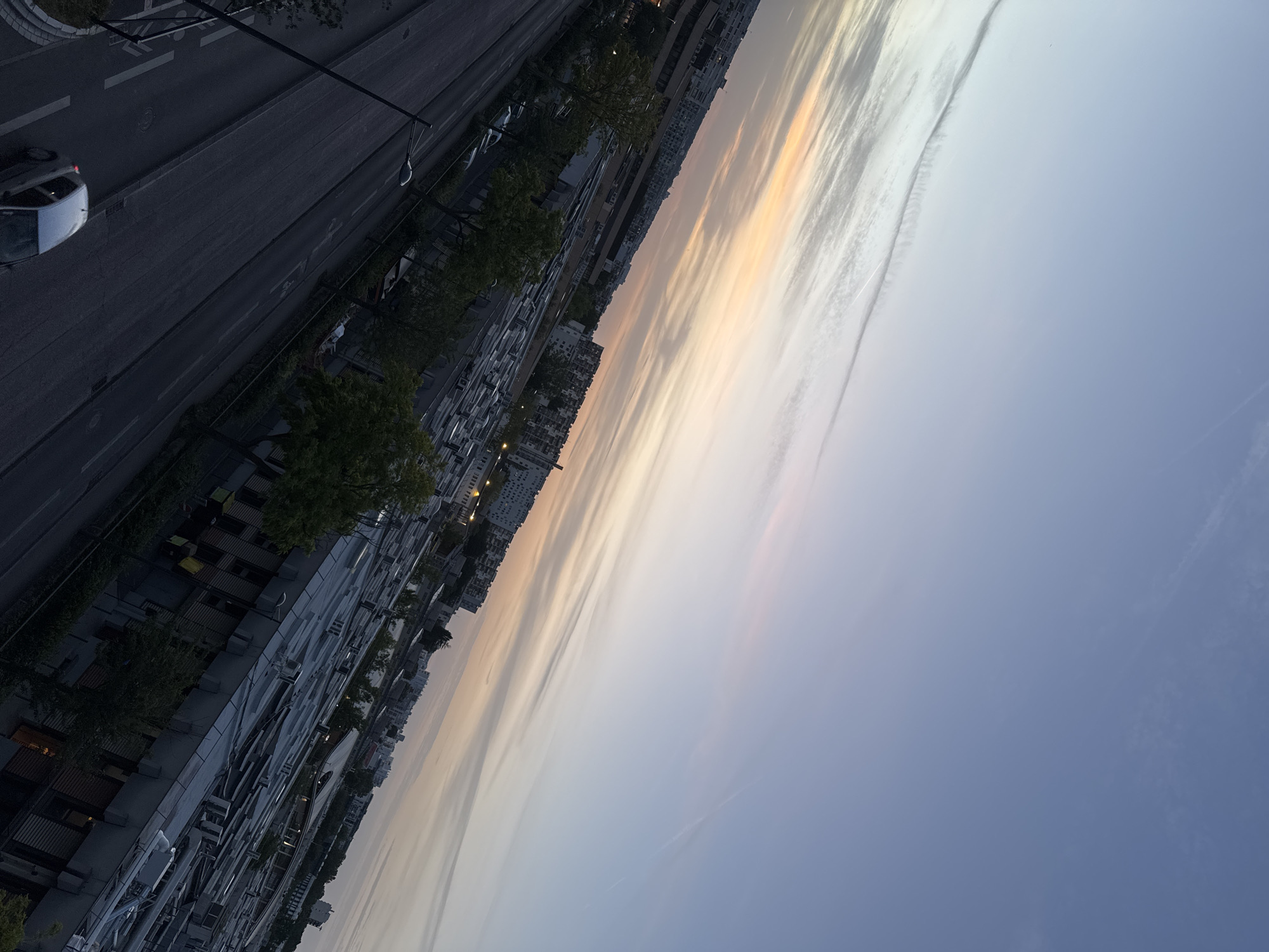 An angled, high-angle shot captures a cityscape at dusk. The sky displays a gradient of soft blues and oranges, with streaks of clouds indicating an approaching sunset or sunrise. Below the sky, buildings of varying heights and architectural styles dominate the mid-ground and background, some appearing as apartment blocks and others as industrial or commercial structures with flat, metallic roofs. Trees with green foliage dot the scene, interspersed between buildings and along the roadside. In the foreground, a dark asphalt road runs diagonally across the frame, marked with white lines. A single silver car is visible on the road, moving away from the viewer, with its brake lights illuminated. Streetlights are present along the road. The overall impression is an urban environment transitioning from day to night.