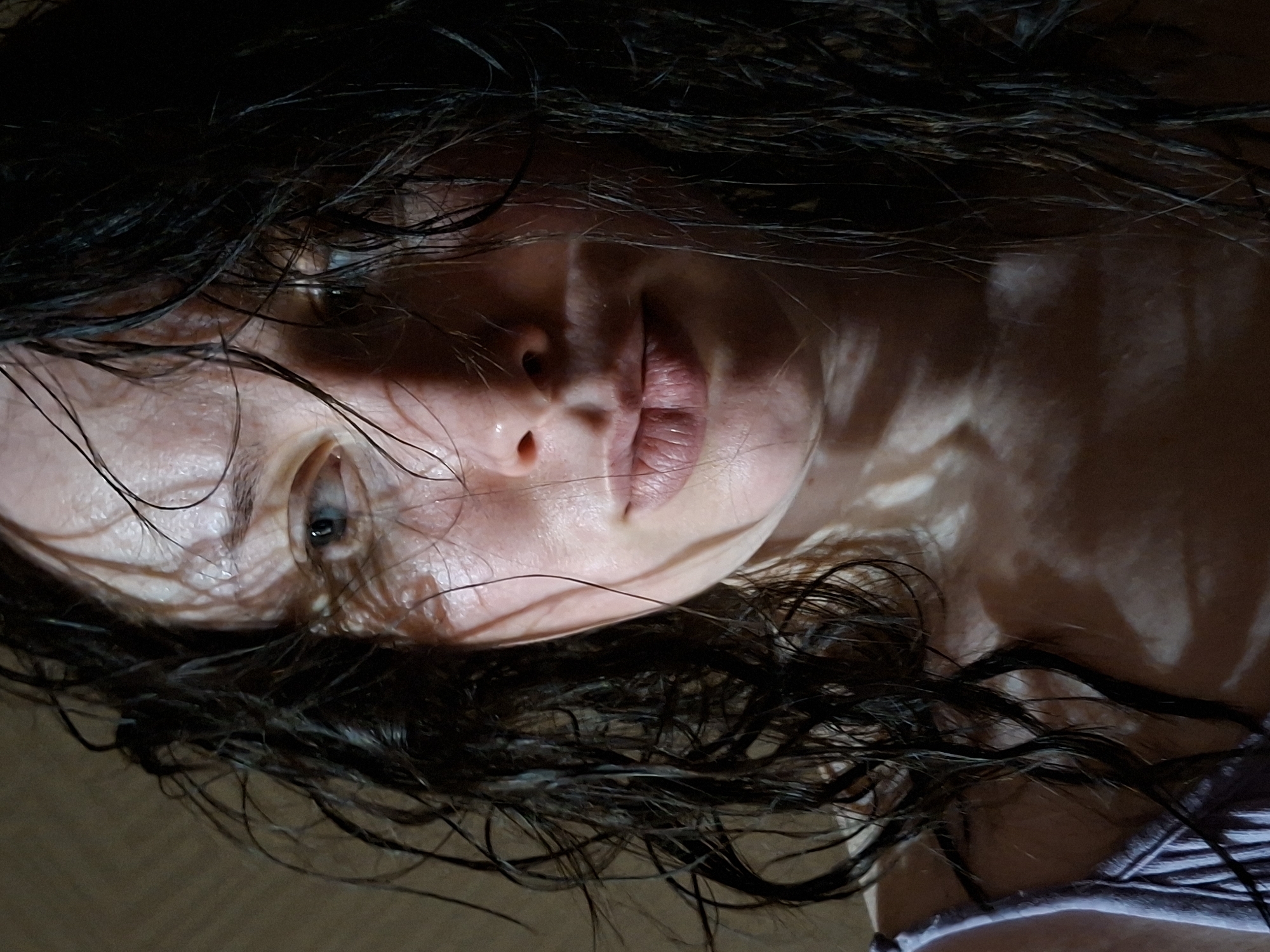 A close-up, tilted perspective shows a person's face and upper chest. They have fair skin, and their hair is dark, wet, and disheveled, framing their face. One eye is visible and appears light in color. The skin has a sheen of moisture, possibly sweat or water. The person's lips are closed, and the lighting is dramatic, with strong shadows and highlights emphasizing the contours of the face. A portion of a purple and white striped garment is visible at the bottom right of the image. Location: Unknown, Unknown.