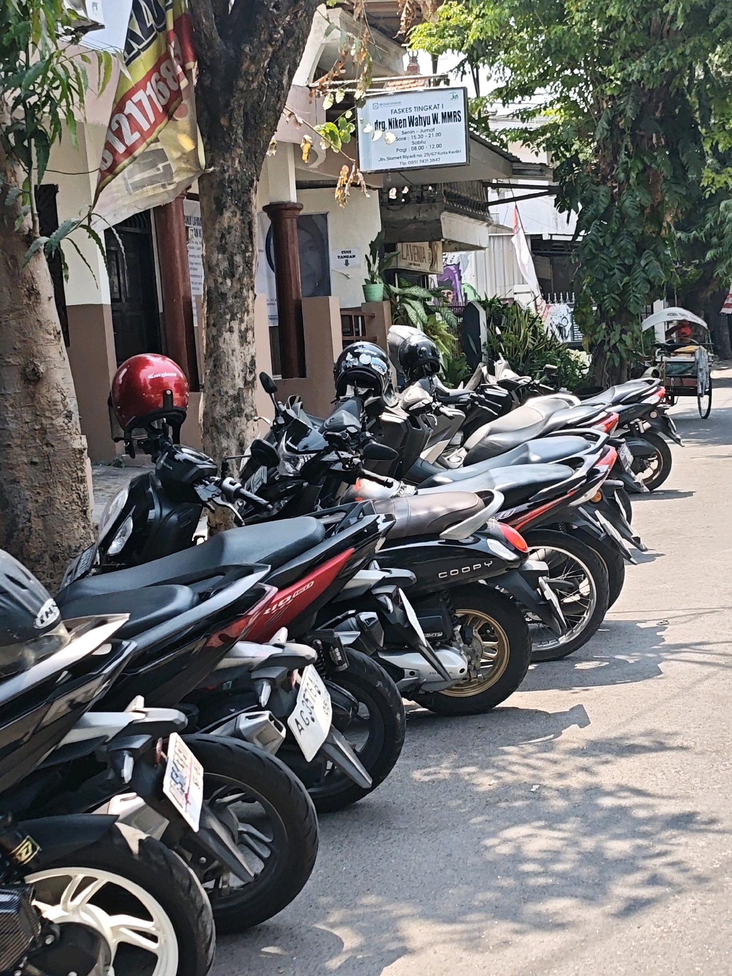 In Kediri City, Indonesia, a row of parked motorcycles lines a sun-dappled street in front of a building. A red helmet rests on the handlebars of a black motorcycle in the foreground. Further down the row, other motorcycles, mostly black and red, are parked. A sign above the building reads "FASKES TINGKAT I drg. Niken Wahyu W. MMRS" and provides operating hours and an address: "Jl. Slamet Riyadi No. 29/67 Kota Kediri". A traditional pedicab with a yellow canopy is visible further down the street. The environment is outdoor, with trees casting shadows on the pavement.
