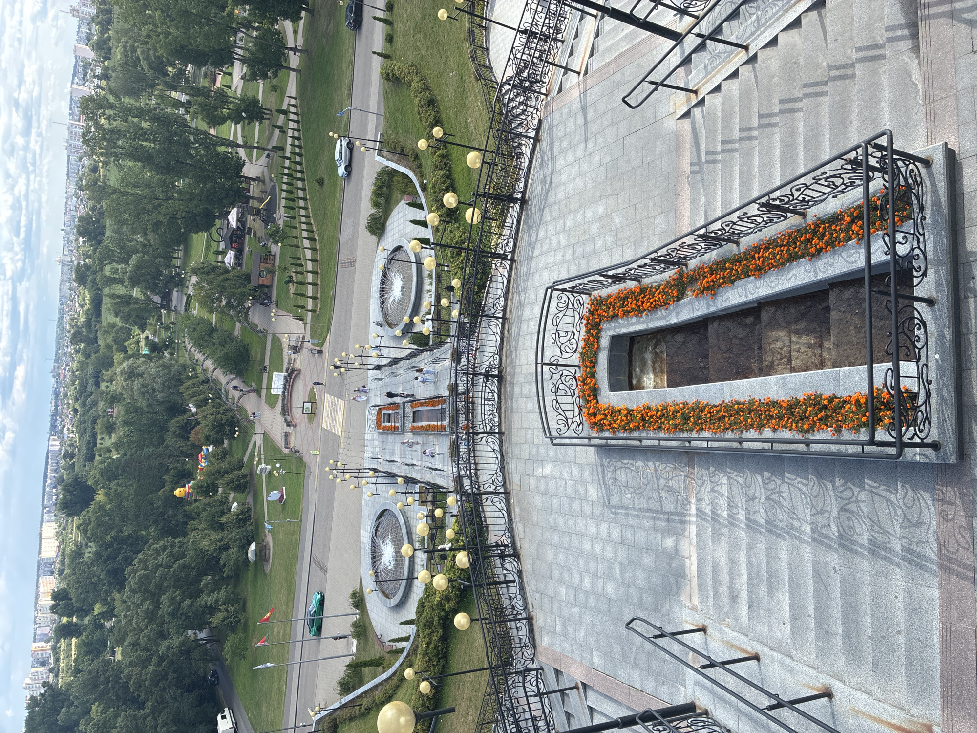 This is a high-angle, medium shot photograph taken on a cloudy day overlooking a park and city landscape in Mahilyow, Belarus. The foreground features a wide set of stone stairs with ornate black metal railings. A long planter box filled with bright orange flowers runs along one side of the stairs. Beyond the stairs, a paved walkway leads to a circular fountain with water spraying upwards. Several people are visible walking on the walkway near the fountain.
In the midground, the park area is characterized by lush green trees, pathways, benches, and decorative lighting fixtures with round, yellow globes. A few small buildings, possibly kiosks or pavilions, are scattered throughout the park. Cars are present on a road bordering the park, including a green car driving on the left side of the road.
The background reveals a cityscape with numerous tall, modern buildings, suggesting an urban environment. The sky is overcast with visible clouds. The overall scene depicts a public park within a city, likely a popular recreational area.