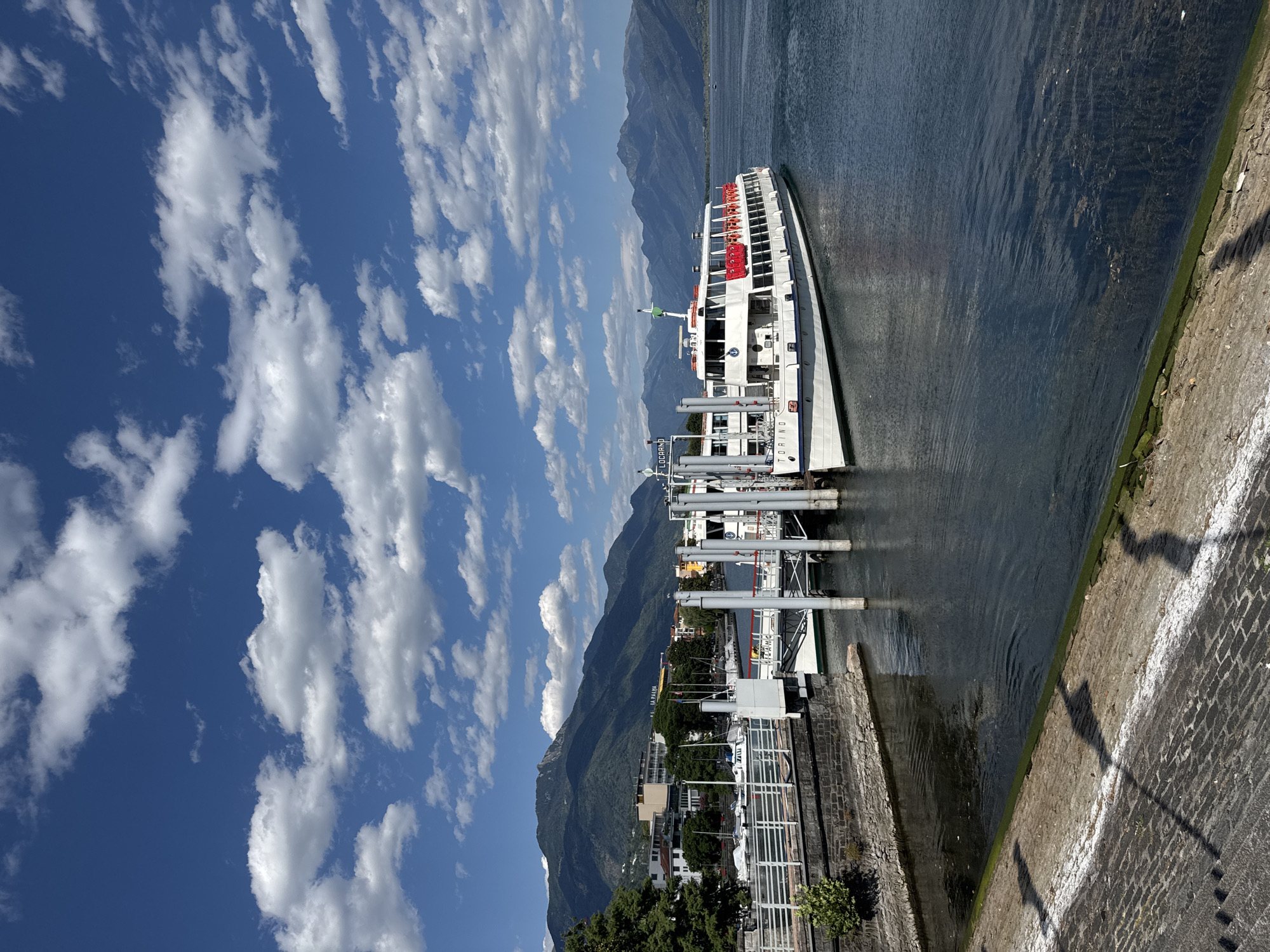 A medium-sized white passenger ferry, named "TORINO" on its hull, is docked at a pier on Lake Maggiore in Locarno, Switzerland. The ferry has a white superstructure with a red trim around its upper deck windows and along the roofline. A white railing with red life preservers is visible on the upper deck. Two other vessels are partially visible behind the "TORINO" at the pier.
The ferry is moored to a substantial gray and white pier with multiple vertical pilings. The waterfront features a stone embankment and a walkway constructed of cobblestones. Greenery, including trees and shrubs, is present along the embankment and the town's shoreline.
In the background, the town of Locarno is nestled against a backdrop of lush green mountains that rise steeply. The sky is a vibrant blue, dotted with scattered white cumulus clouds. The water of Lake Maggiore is a deep blue-green, with gentle ripples on its surface. The overall scene depicts a sunny day in a picturesque lakeside town.