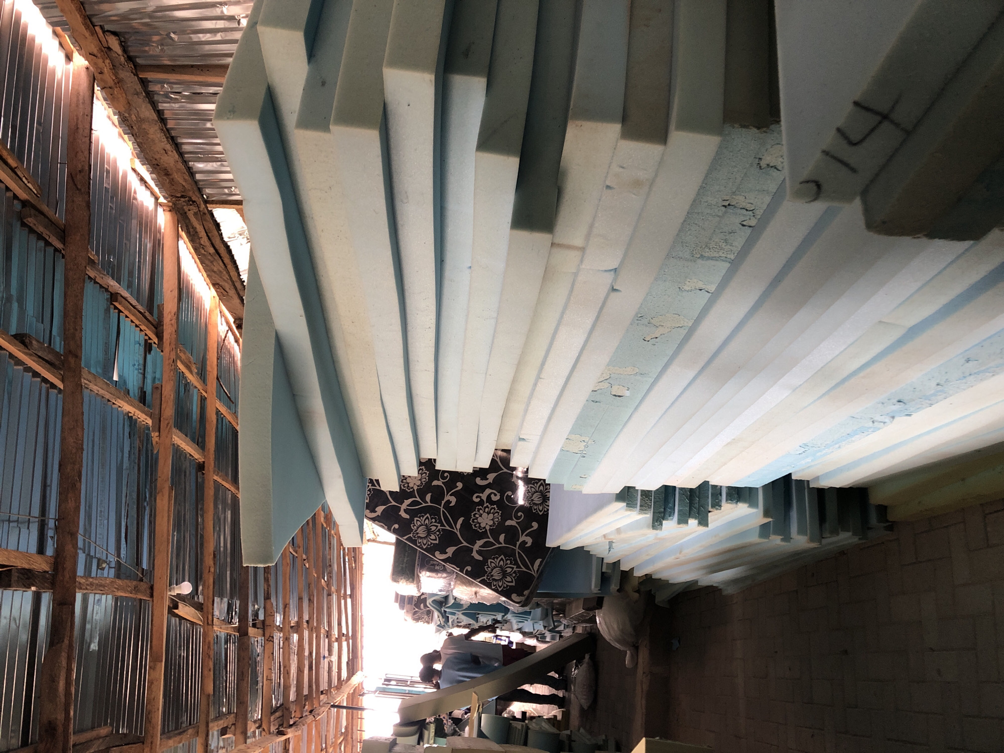 The image shows the interior of a building in Ado, Nigeria. On the left, a corrugated metal wall with wooden supports is visible, illuminated by daylight streaming through gaps. Stacks of rectangular foam blocks, primarily white and light blue, are piled high and extend across the frame. Some blocks have markings, possibly numbers. A sofa with a black and white floral pattern is situated between the foam stacks. In the distance, glimpses of machinery and a person in light clothing are visible in the brightly lit area. The perspective is rotated, making it appear as though the foam stacks are hanging overhead.