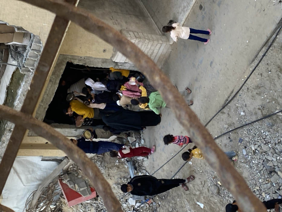 The image, captured in Khan Yunis, Palestinian Territory, presents an elevated view through a rusty metal grate overlooking an outdoor scene. Approximately 20-30 individuals, predominantly children and several adults, are gathered near and emerging from a dark, rectangular doorway of a partially damaged building. The left side of the scene is covered in substantial construction rubble, including fragmented concrete, rebar, and debris. Multiple individuals, including a man in a yellow shirt and another in a dark blue shirt, are carrying metal pots and bowls, suggesting food distribution or collection. A woman in a black hijab and abaya stands centrally near the doorway, surrounded by children. The ground consists of packed dirt and loose gravel, with two dark cables extending across it. A young man in a black tracksuit stands on the left, while a young girl in a light-colored top and blue pants stands on the far right. The building's entrance is set within a concrete wall, with additional structural damage visible above and to the left of the doorway.