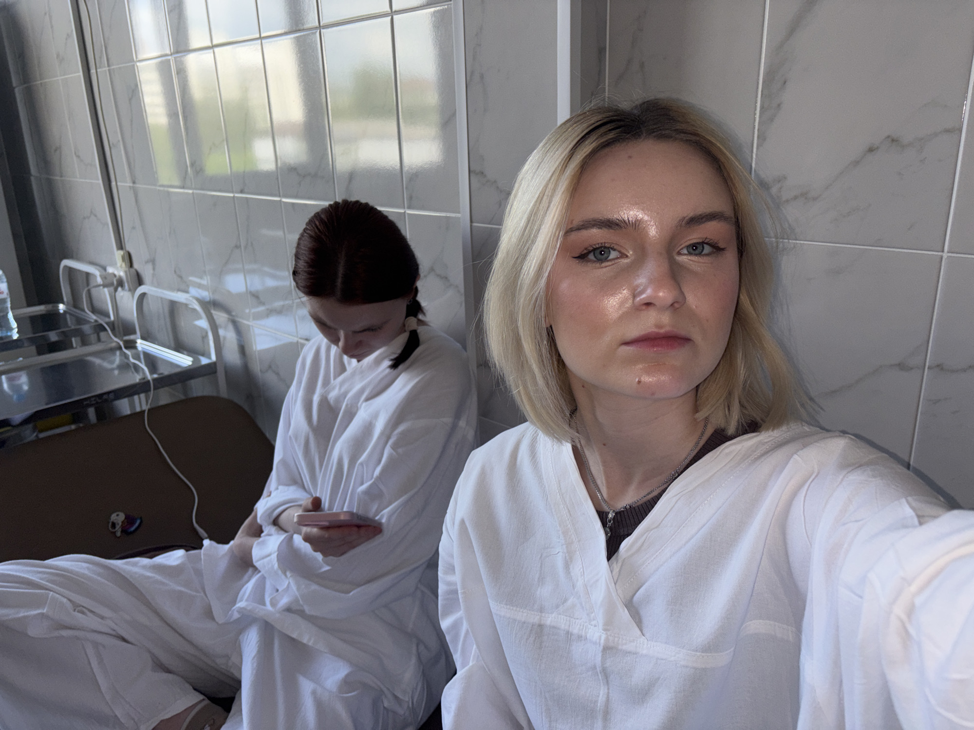 Two young women are captured in what appears to be a medical facility, possibly in Saint Petersburg, Russia. The woman in the foreground, with blonde hair, is taking a selfie, looking directly at the camera with a neutral expression. She wears a white, loose-fitting garment, resembling a hospital gown or patient attire, over a dark top and a silver chain necklace.
Behind her, to the left, a second woman with dark hair (possibly red or auburn), styled in a braid or ponytail, is seated and looking down, engrossed in a pink smartphone held in her hands. She is also dressed in a similar white garment, covering her body and legs.
The setting is characterized by light gray tiled walls with subtle marble-like patterns. Reflections in the window behind the subjects suggest an urban environment with buildings visible outside. Medical equipment is present, including a stainless steel cart or table with a water bottle and what might be a charging cable or IV line. A brown surface, possibly a medical bed or examination table, is also visible.
The scene conveys a quiet moment, possibly during a waiting period or recovery in a clinic or hospital, during daytime, given the bright natural light. No distinct, legible text is visible on any objects or surfaces.